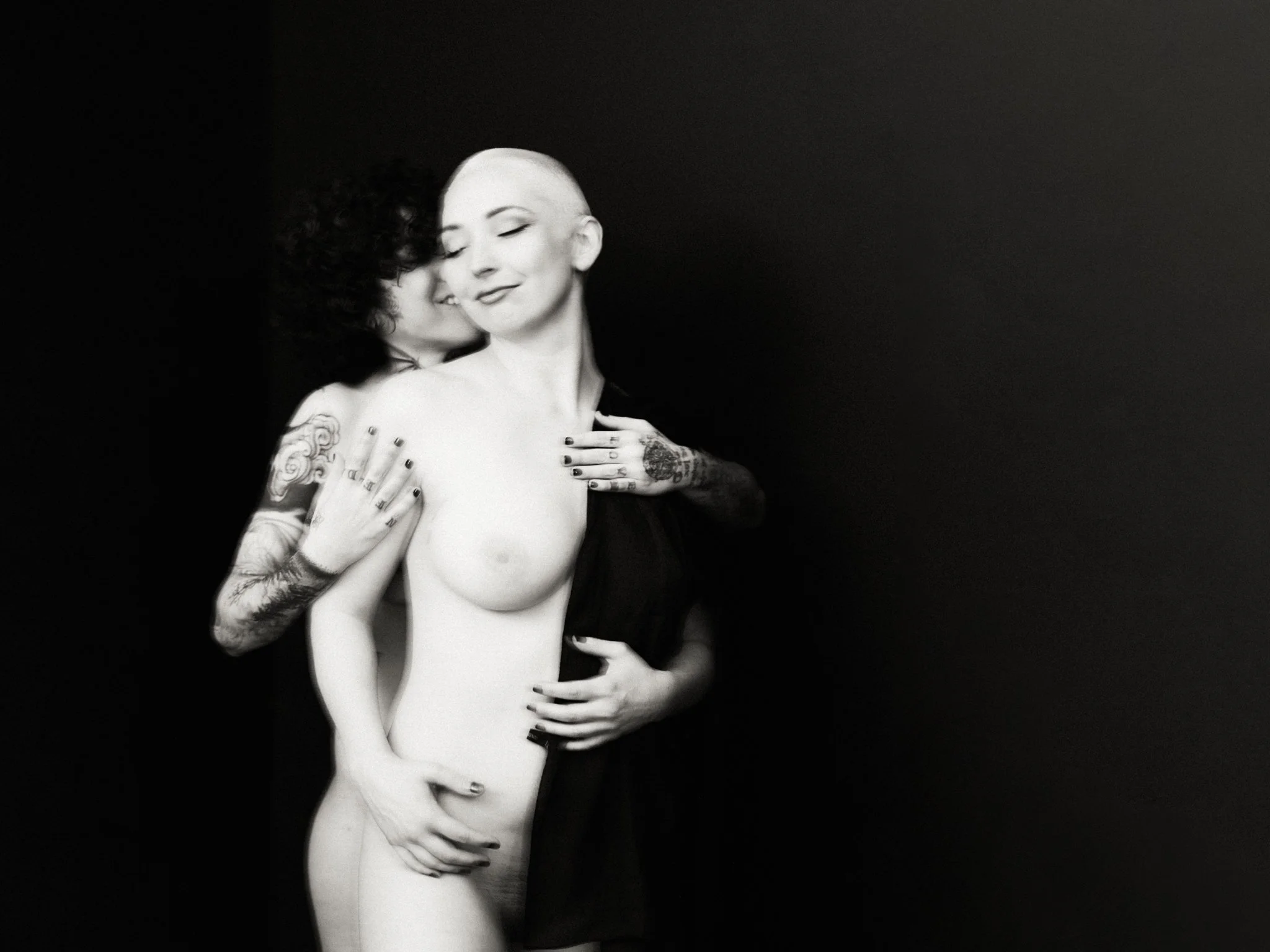 Black and white photograph of two women embracing, one with dark curly hair and the other with a shaved head, tattoos, and a faint smile, against a plain dark background.