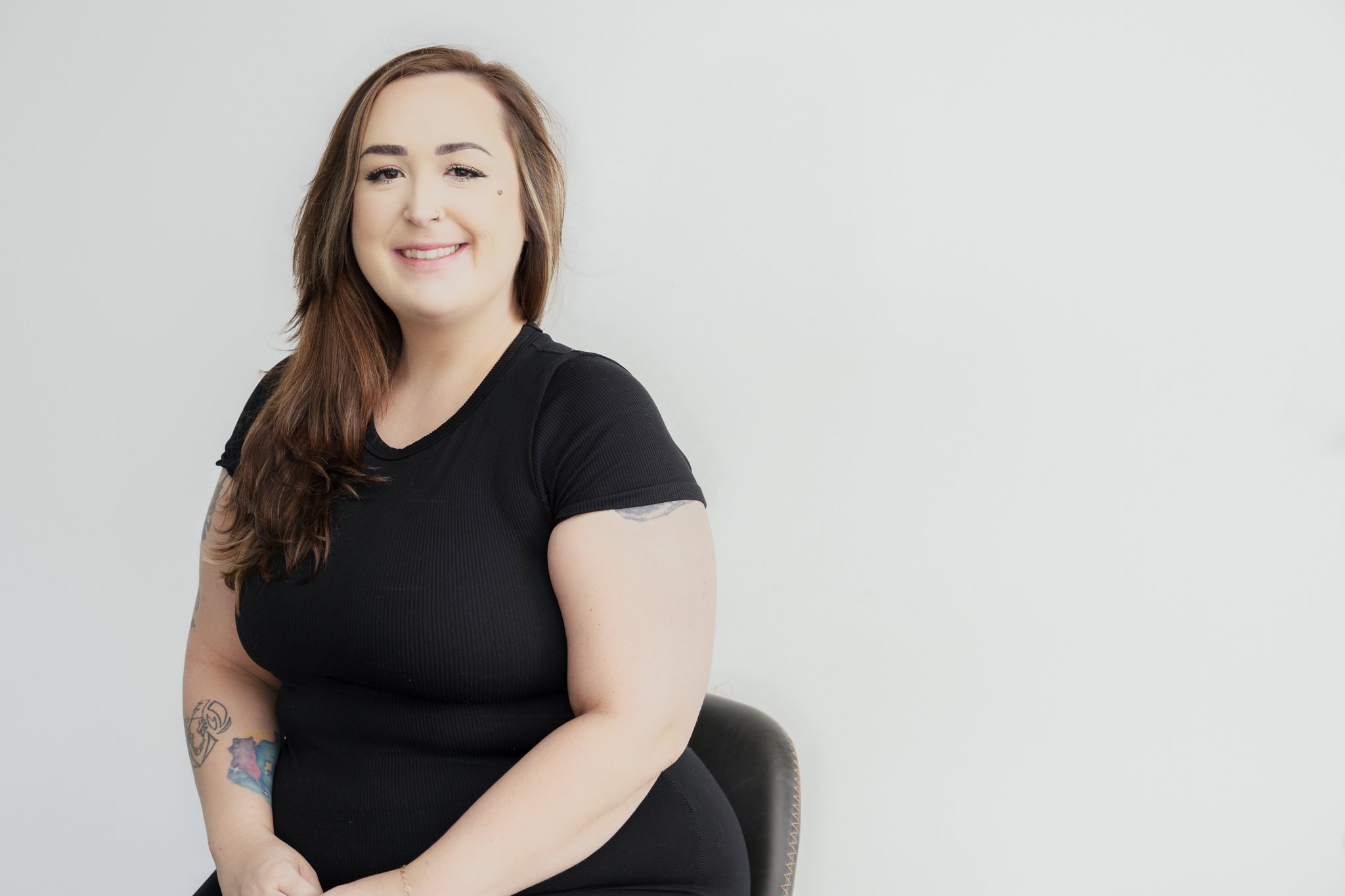 A smiling woman with shoulder-length brown hair, sitting on a black chair against a plain white background, wearing a black t-shirt with tattoos on her arm.