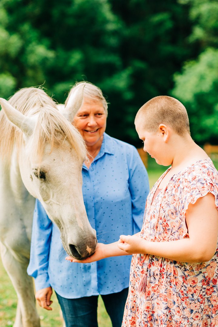 A young girl and an older woman, possibly her grandmother, are outdoors with a white horse. The woman is smiling as the girl gently touches the horse's nose, and they are surrounded by green trees in the background.