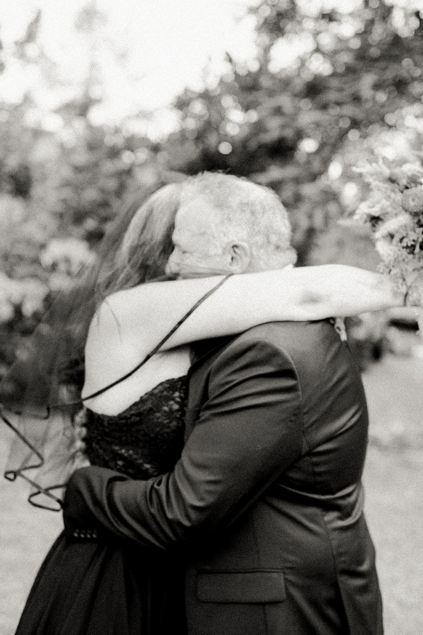 A black and white photo of a couple hugging passionately outdoors, with trees and foliage in the background.