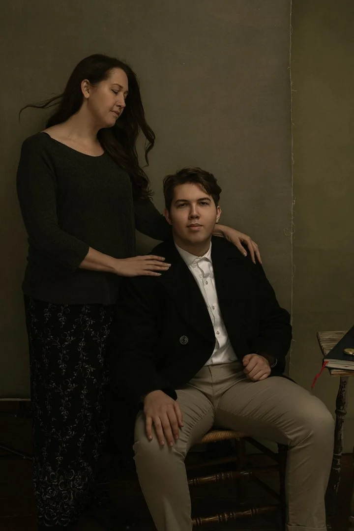 A woman with long dark hair wearing a black top and patterned pants standing beside a seated young man in a black blazer, white shirt, and beige pants, with a plain wall background.