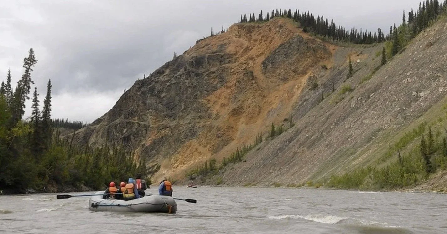 People in life jackets paddling a gray inflatable boat on a river surrounded by mountains with trees.