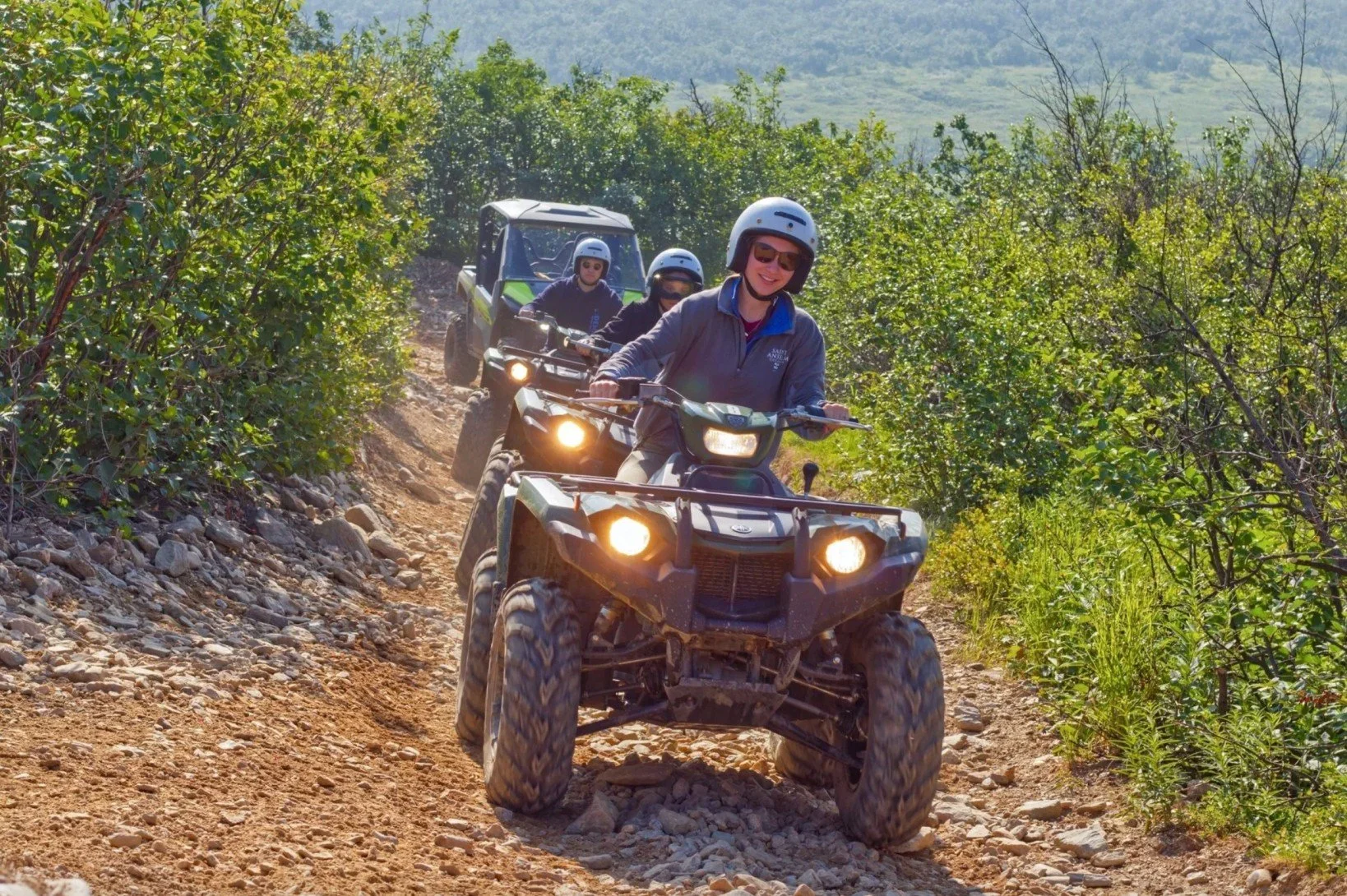 People riding all-terrain vehicles on a dirt trail through a green, bushy landscape with hills in the background.