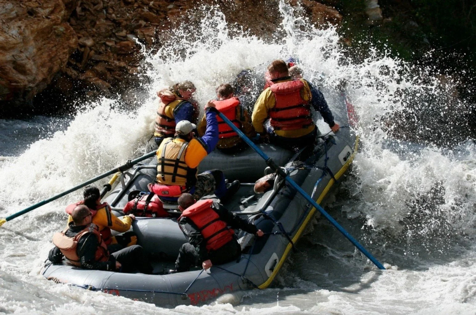 A group of people in a raft navigating white water rapids during a rafting adventure, wearing life jackets and holding paddles.