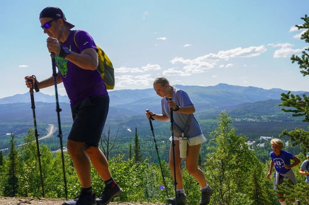 Group of people hiking on a trail through a lush forest with mountains in the background under a partly cloudy sky.