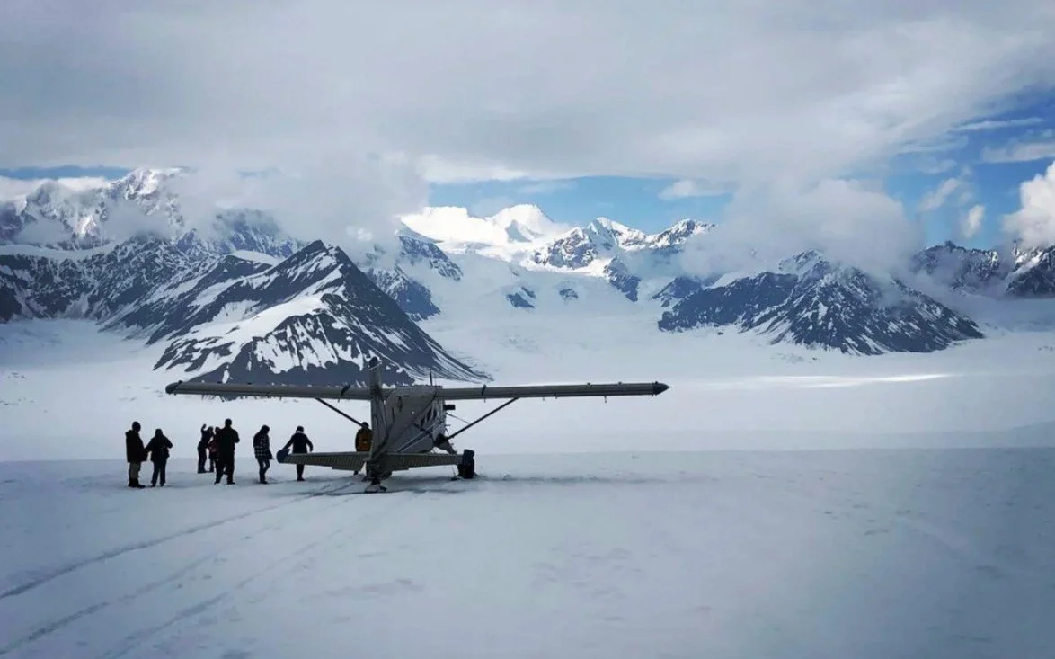 Scientists preparing a drone on snow in a mountainous, polar region with snow-covered peaks and clouds overhead.