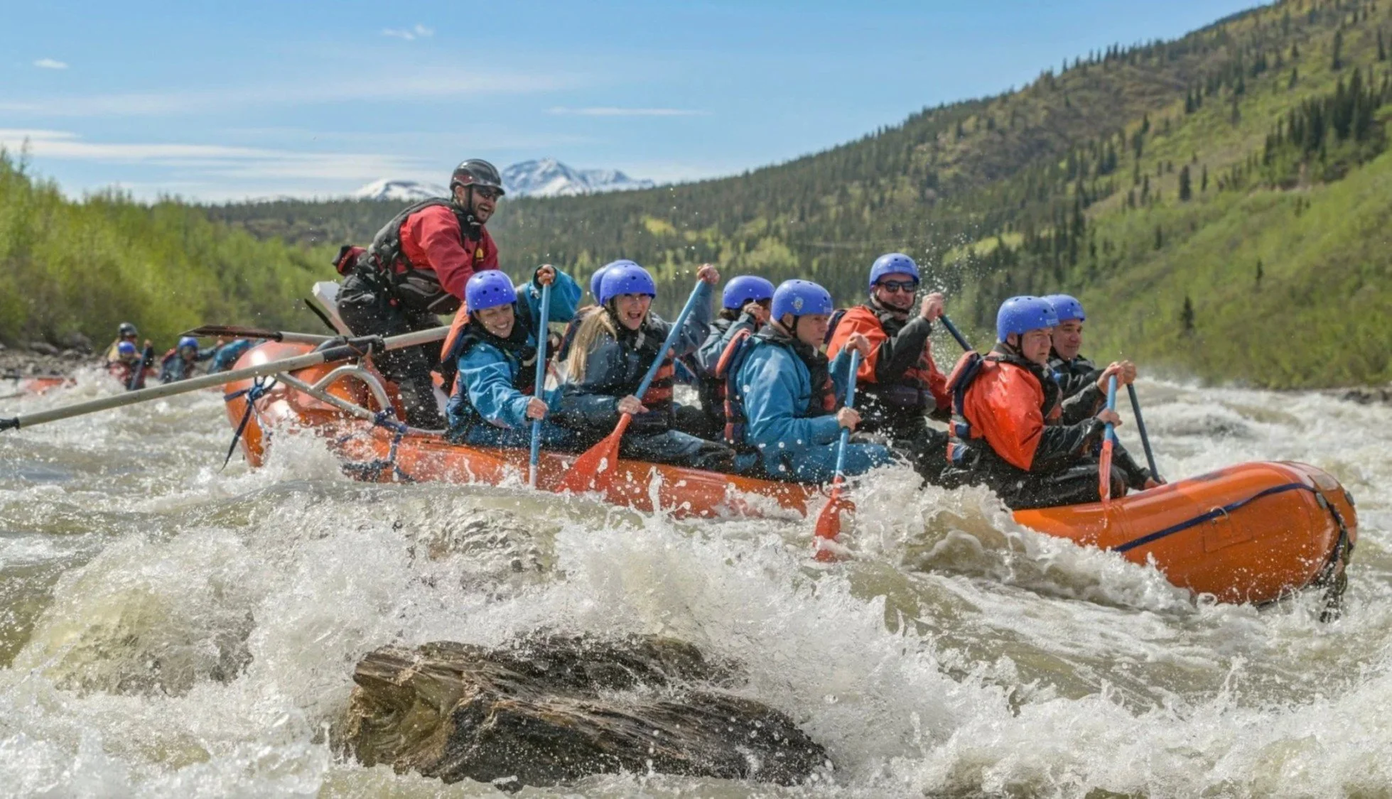 A group of people wearing life jackets and helmets white water rafting on a river with a mountainous landscape in the background.
