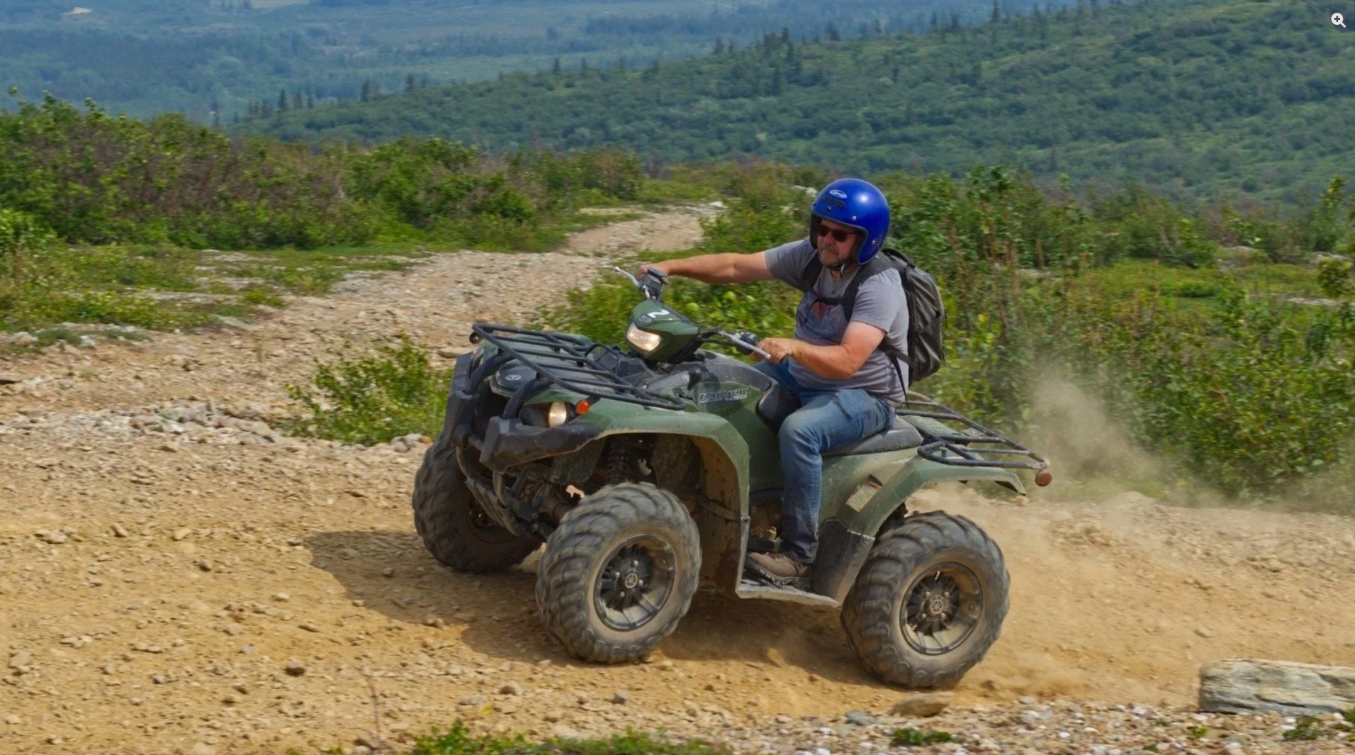 A man riding an all-terrain vehicle (ATV) on a dirt trail in a mountainous landscape, wearing a blue helmet, sunglasses, a gray shirt, blue jeans, and carrying a black backpack.