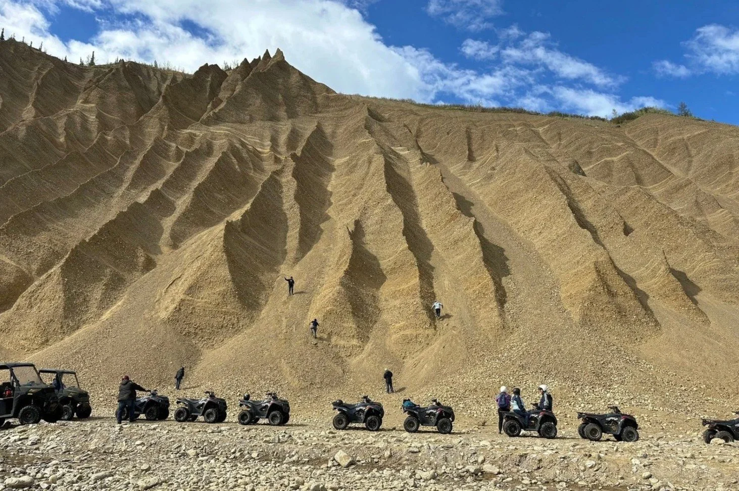 People riding and parking all-terrain vehicles (ATVs) at the base of a large, steep, and rugged cliff or mountain with layered rock formations and a partially cloudy sky.