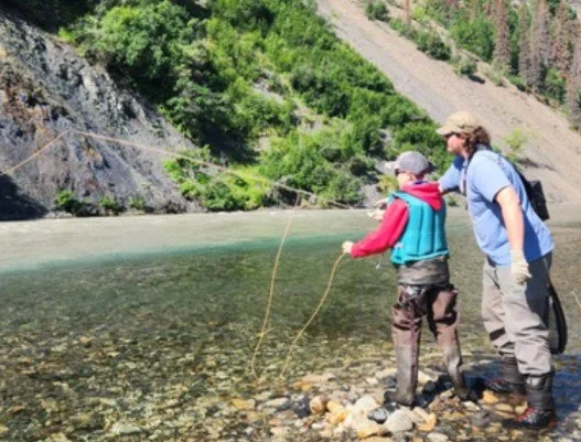 A man and a boy fishing in a river by a rocky shoreline surrounded by trees and steep hillside.