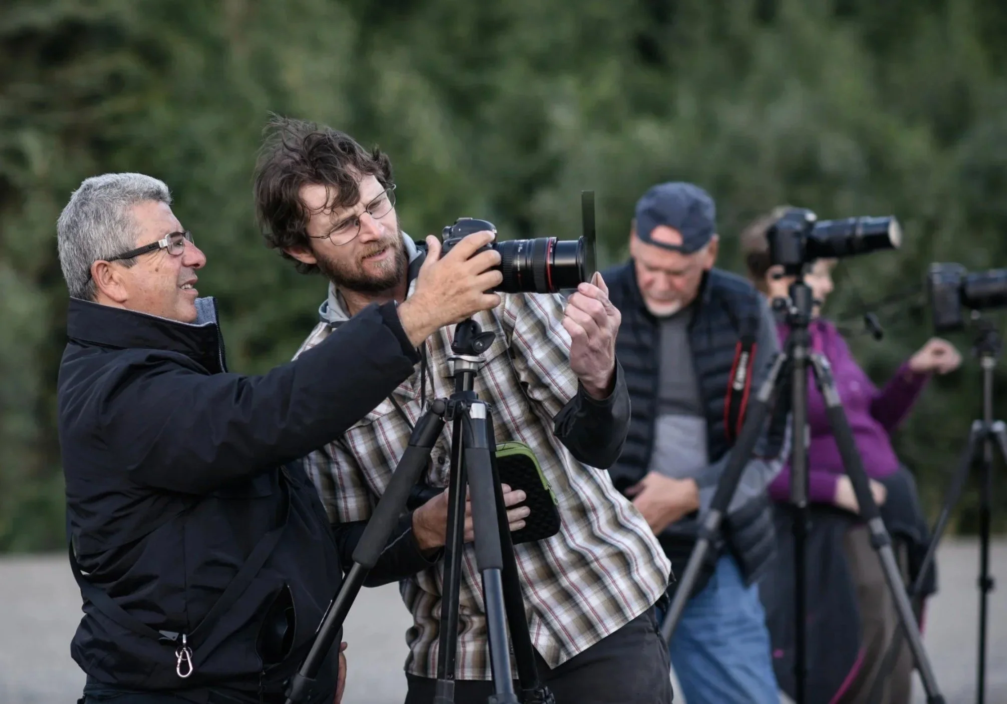 Group of people outdoors with cameras, one man adjusting his camera on a tripod, others using handheld cameras, in a natural setting.