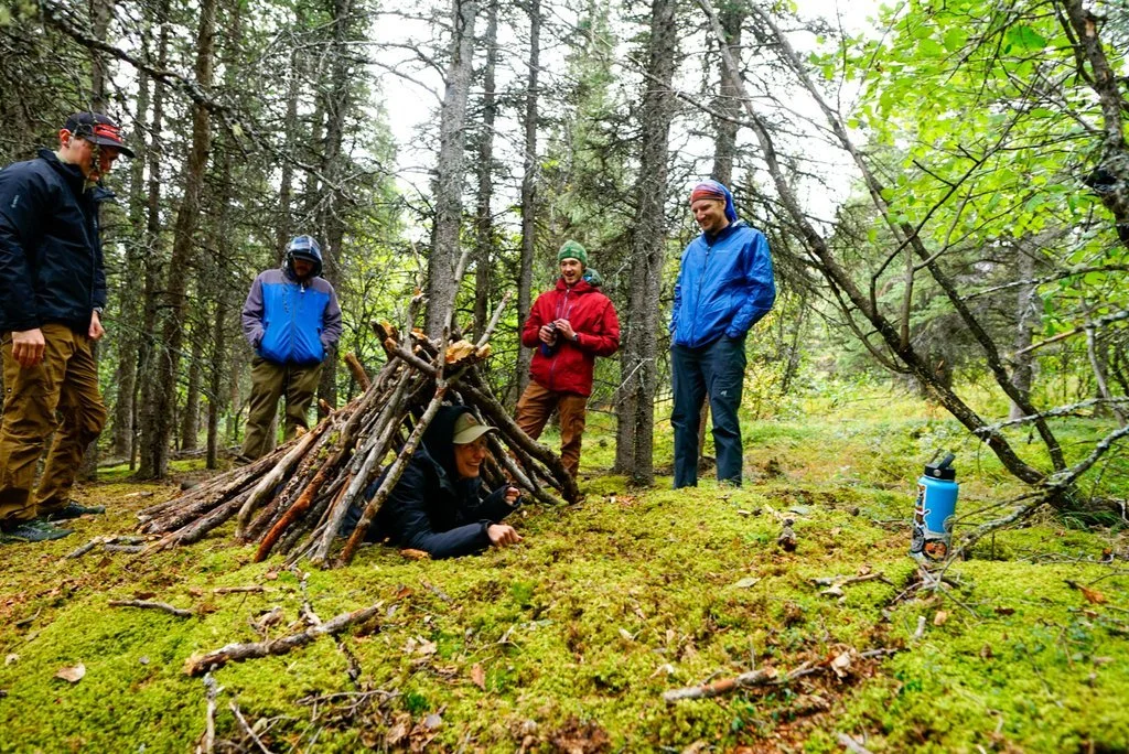 Five people in outdoor clothing gathered around a small shelter made of sticks in a forest, with one person lying inside it and four standing nearby, surrounded by trees and greenery.