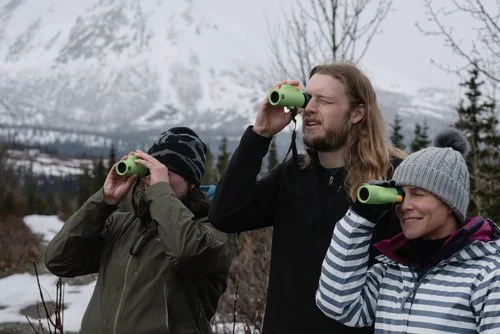 Three people outdoors in a snowy landscape looking through binoculars.