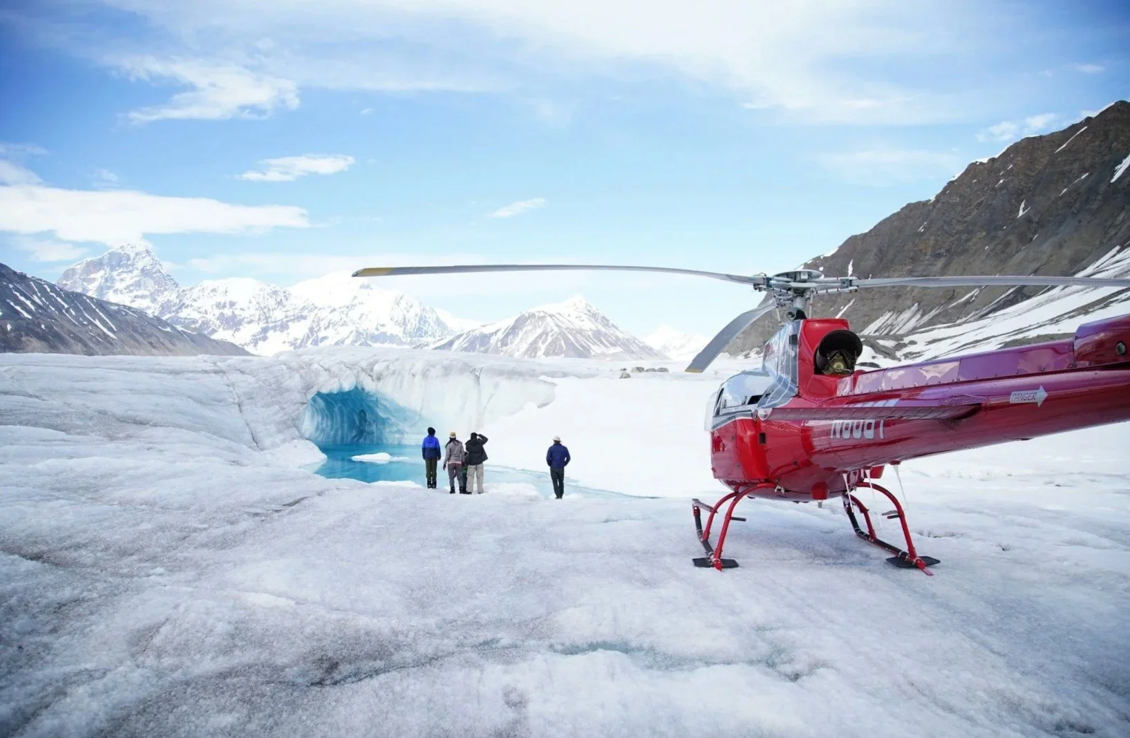A red helicopter parked on snow and ice in a snowy mountainous landscape with four people standing near a glacier's ice cave entrance and snow-capped mountains in the background.