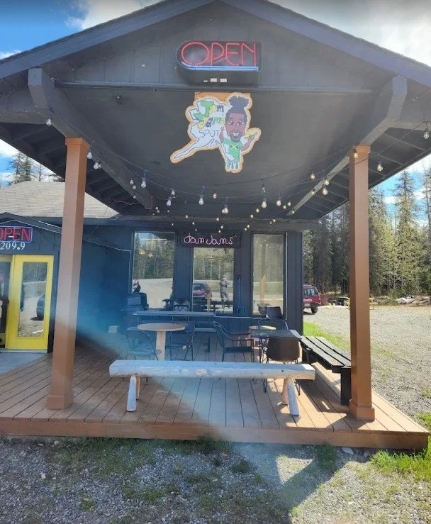 Front porch of a small building with an 'OPEN' neon sign, string lights, a colorful character sign hanging from the ceiling, a large window with seating inside, and outdoor tables and chairs outside.