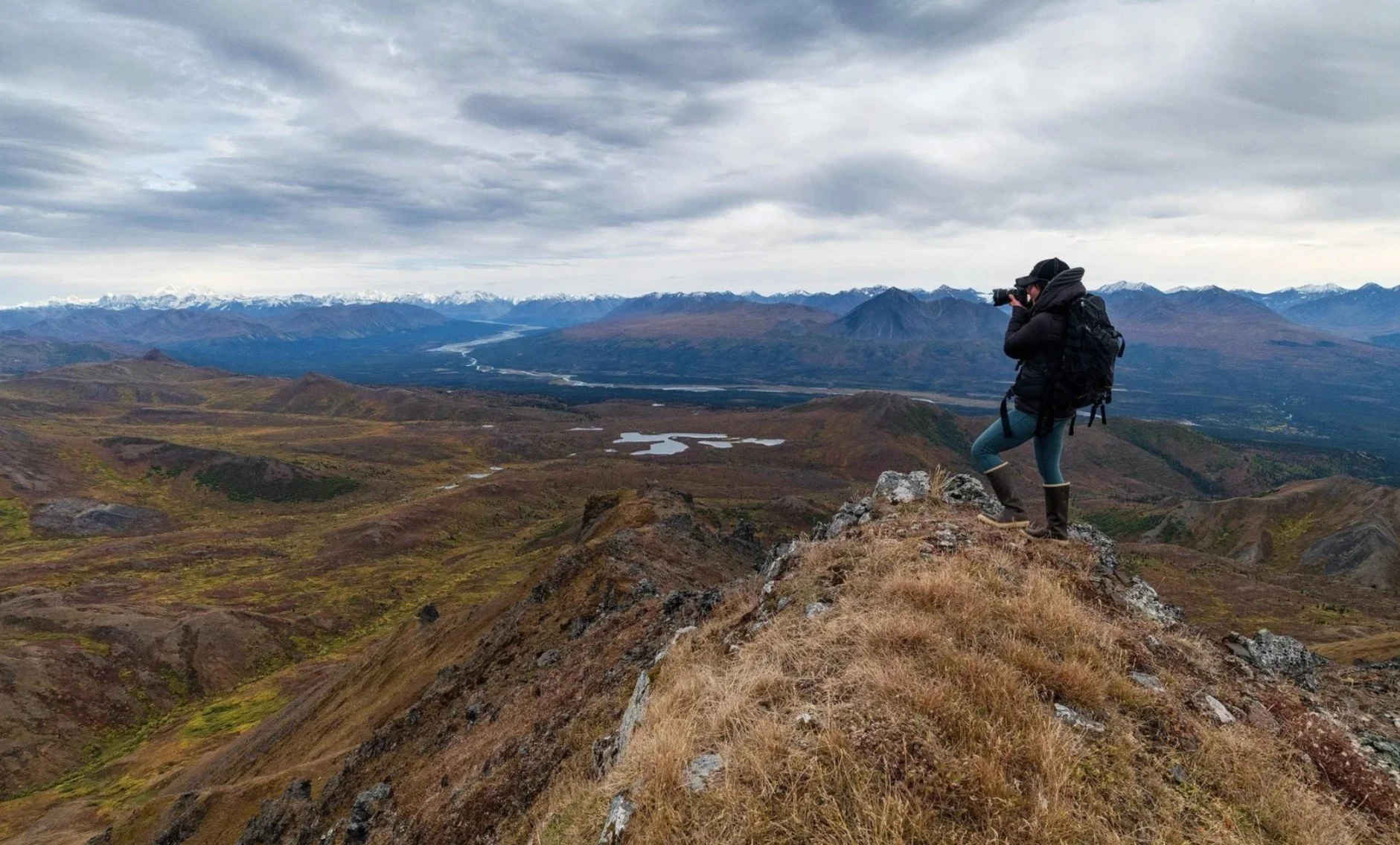 A person wearing outdoor gear, including a backpack, stands on a rocky hilltop taking a photograph of a vast mountainous landscape under a cloudy sky.