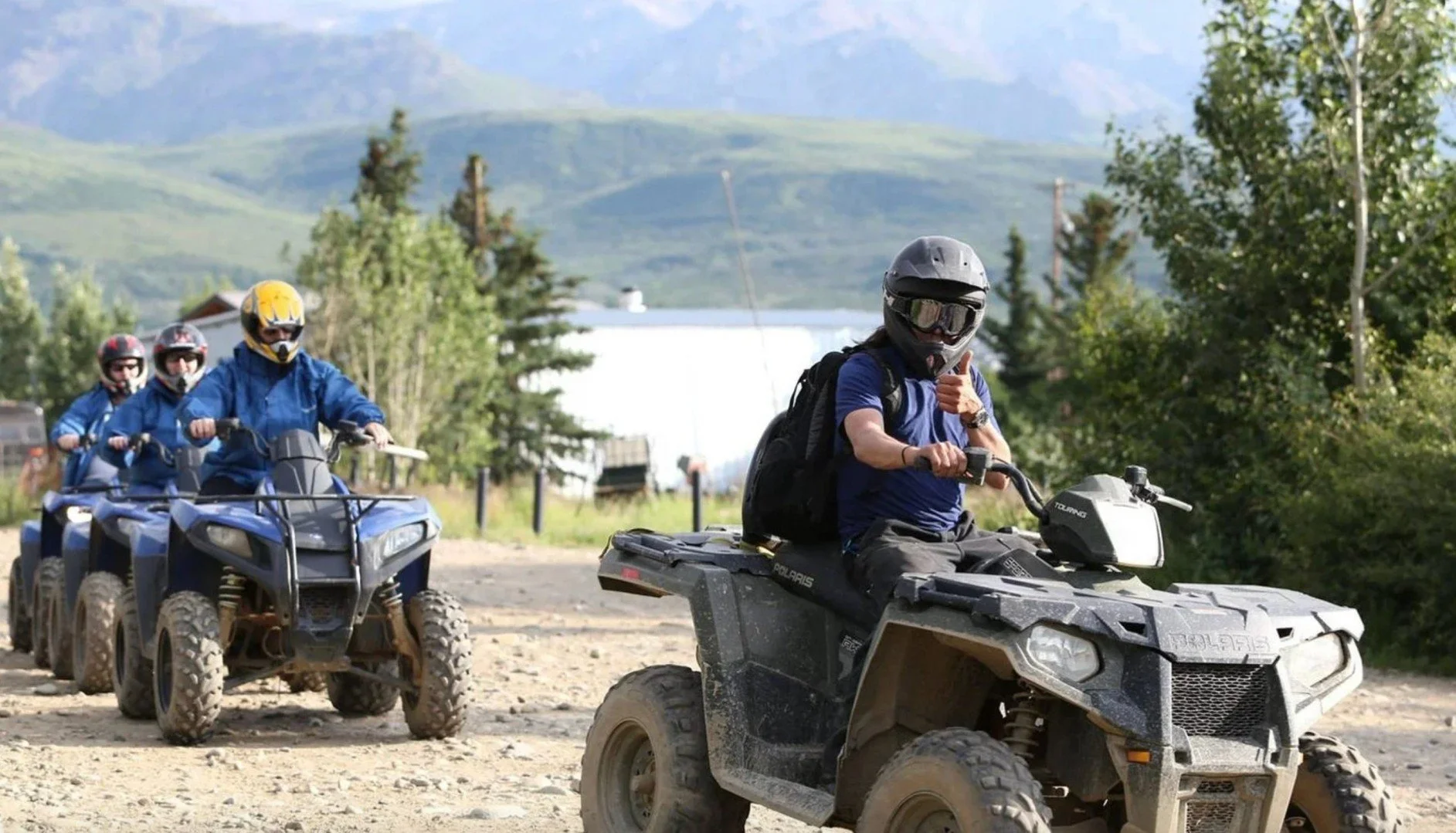 Group of four ATV riders in helmets and protective gear, riding on a dirt path with trees and mountains in the background, the person in front giving a thumbs-up.