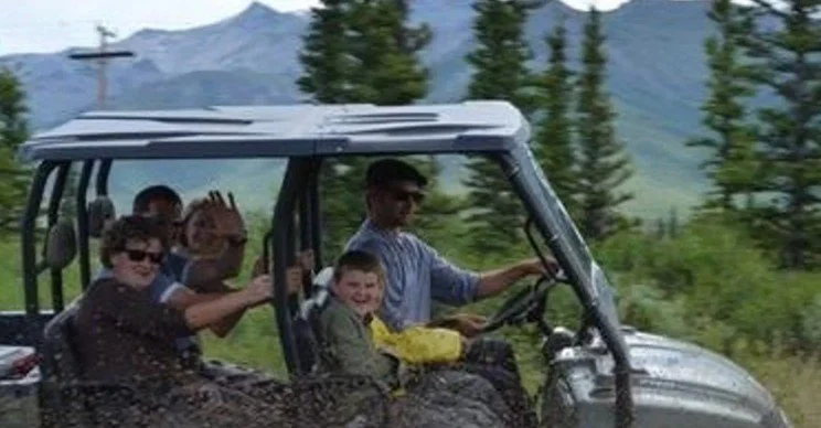 Four children and one adult riding in an off-road vehicle through a forested area with mountains in the background.