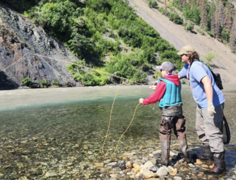 A woman and a boy fishing by a river in a mountainous, forested area.