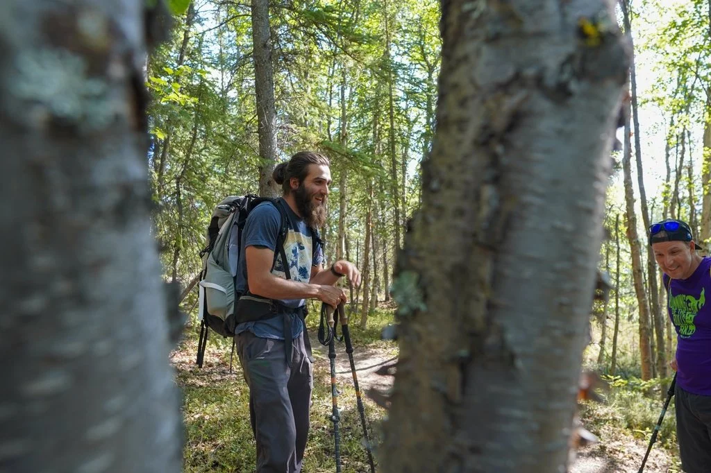 Two men are hiking in a forest, smiling and talking to each other, with the photo taken through the gap of two trees. One has a beard, long hair tied back, a backpack, and hiking poles. The other wears sunglasses and a purple t-shirt with a green graphic.