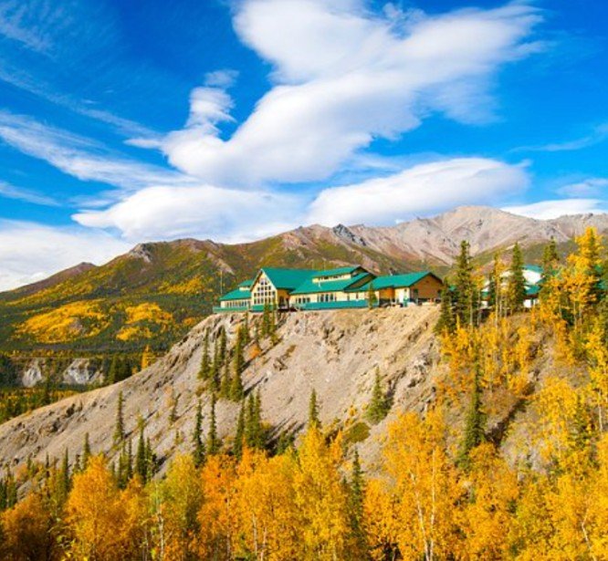 A mountain lodge with green roofs situated on a rocky hillside, surrounded by autumn-colored trees under a partly cloudy sky.