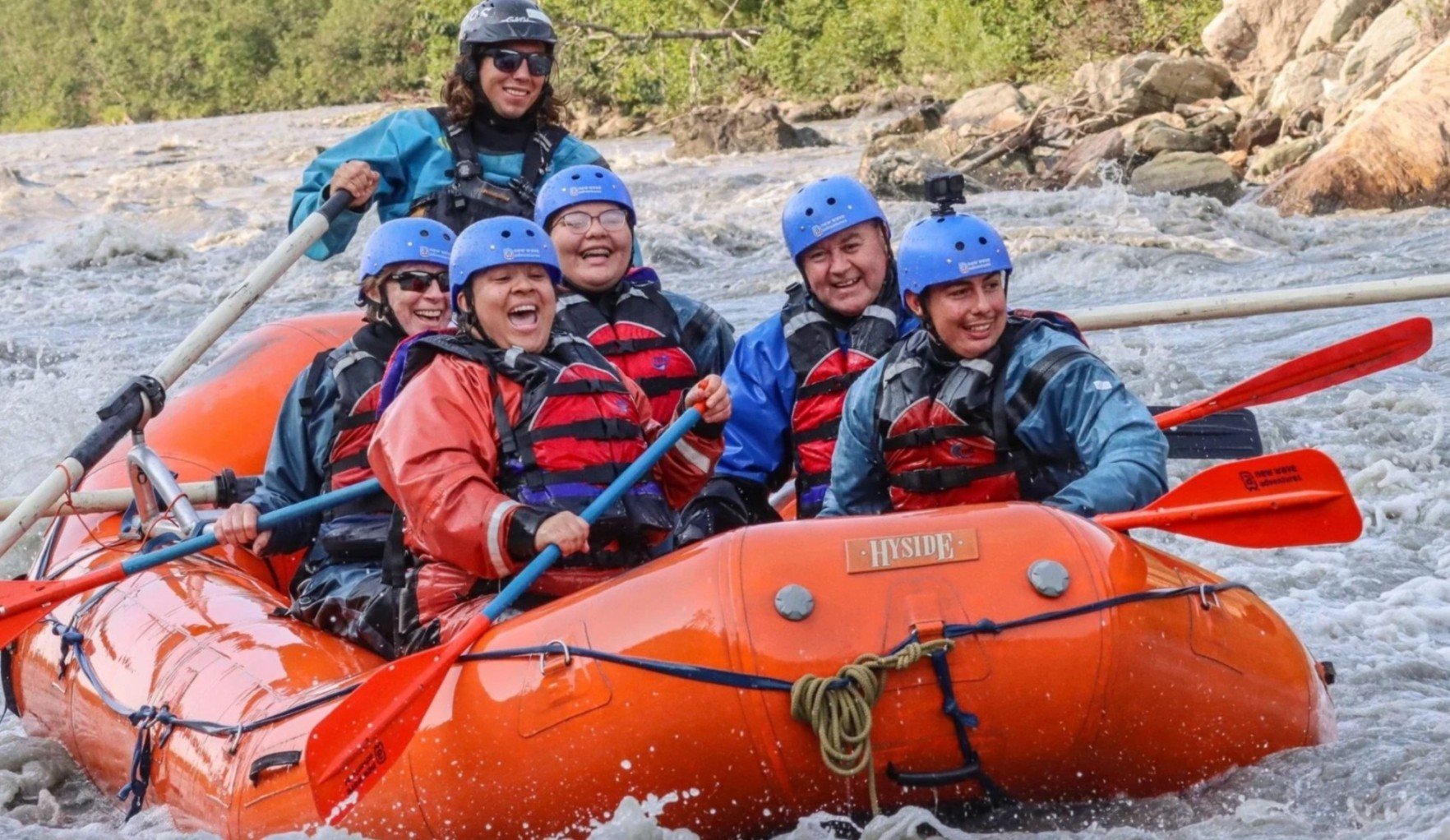 A group of six people, dressed in rain gear and helmets, whitewater rafting on a river.