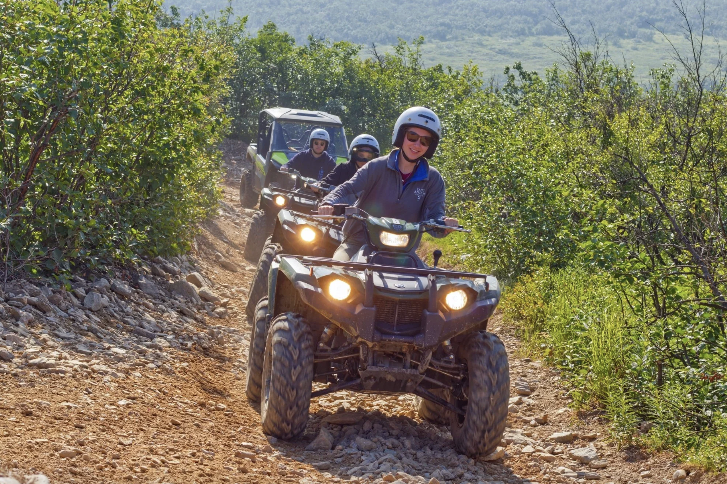 Group of four people riding all-terrain vehicles on a dirt trail surrounded by green bushes and trees.