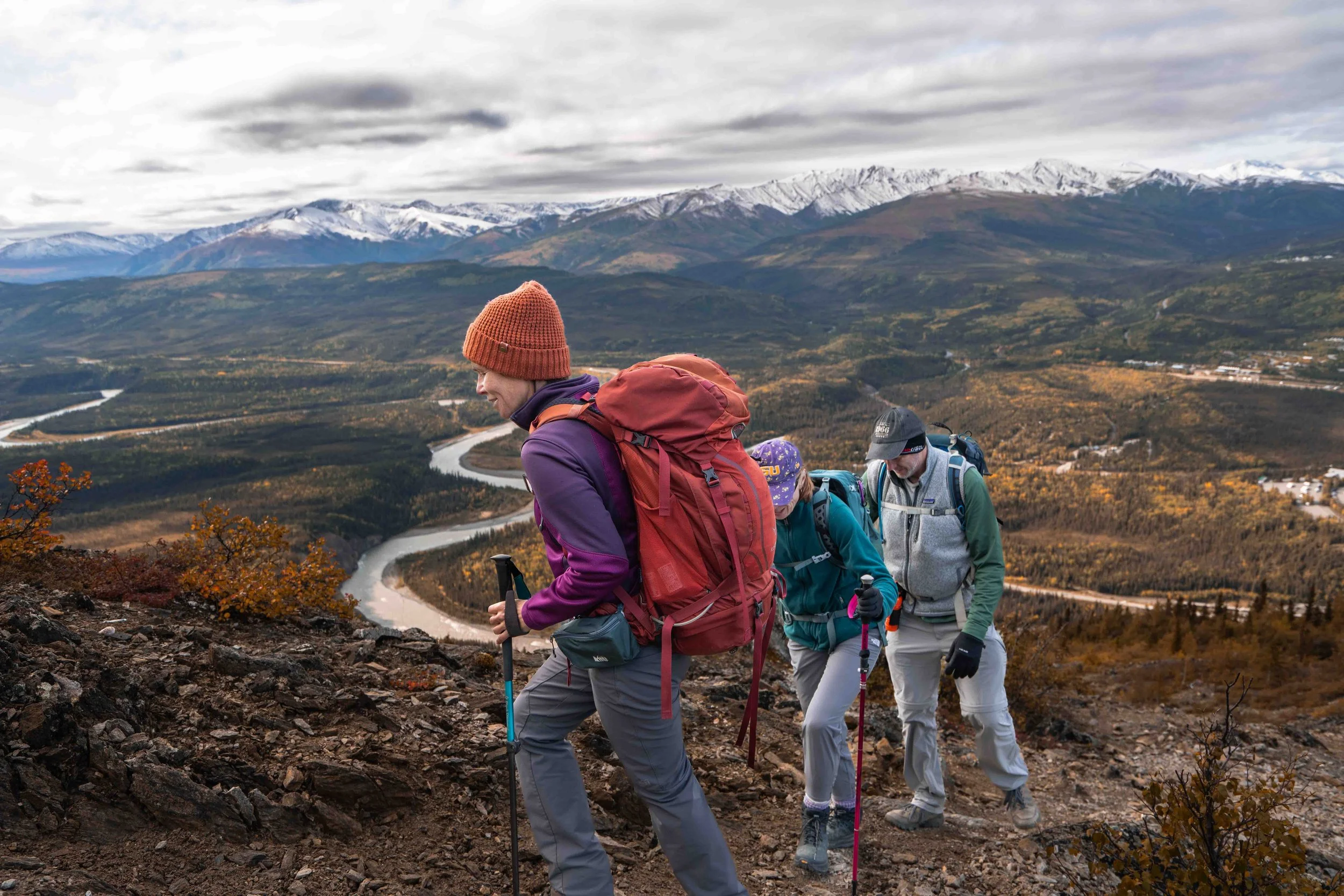 Four hikers wearing backpacks and outdoor clothing ascending a mountain trail with a river winding through a valley and snow-capped mountains in the background.