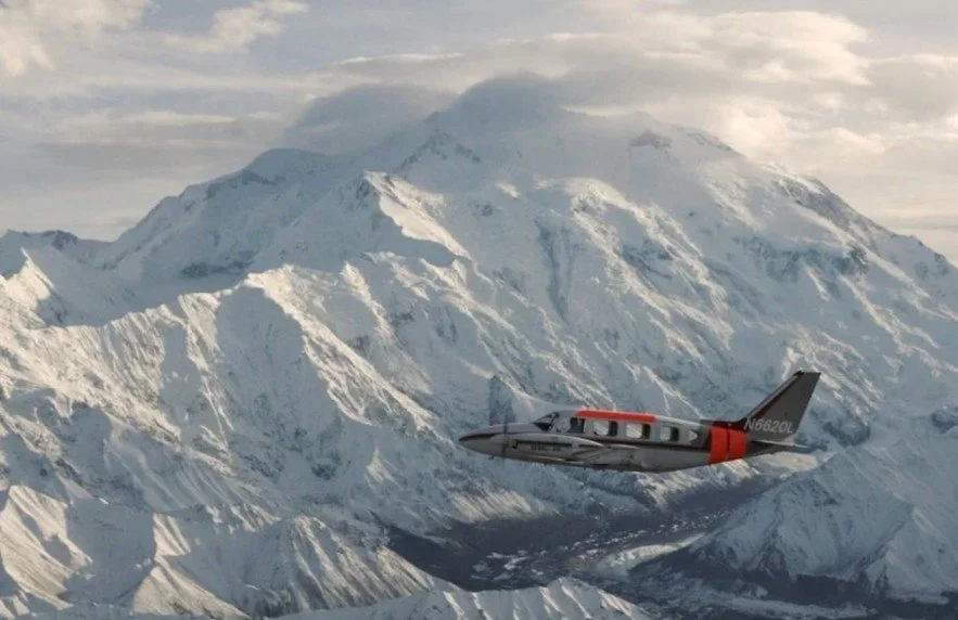An airplane flying over snow-covered mountains with a cloudy sky.