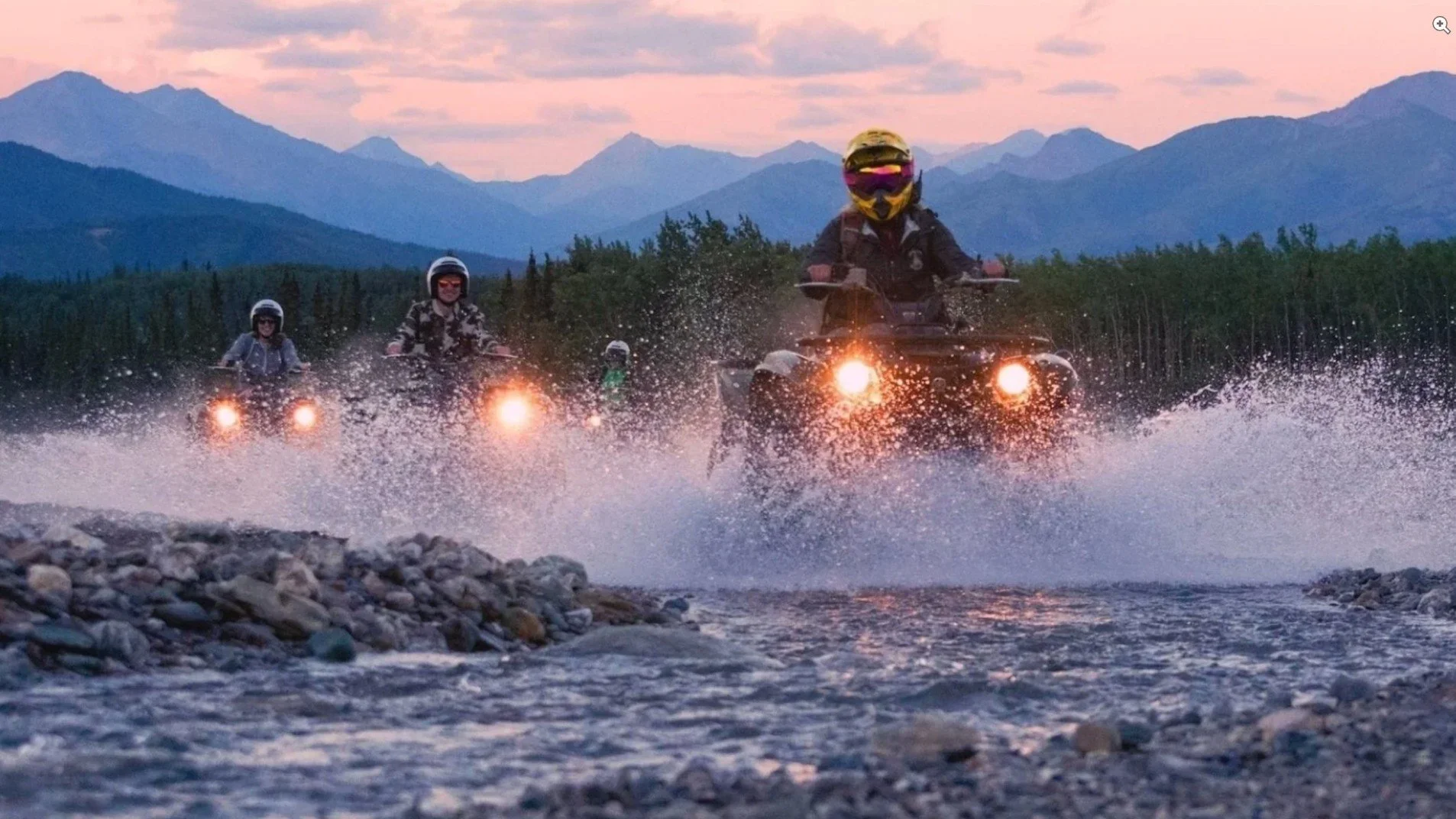 Four people riding all-terrain vehicles (ATVs) through a shallow river at dusk, with mountain ranges in the background and water splashing around the vehicles.