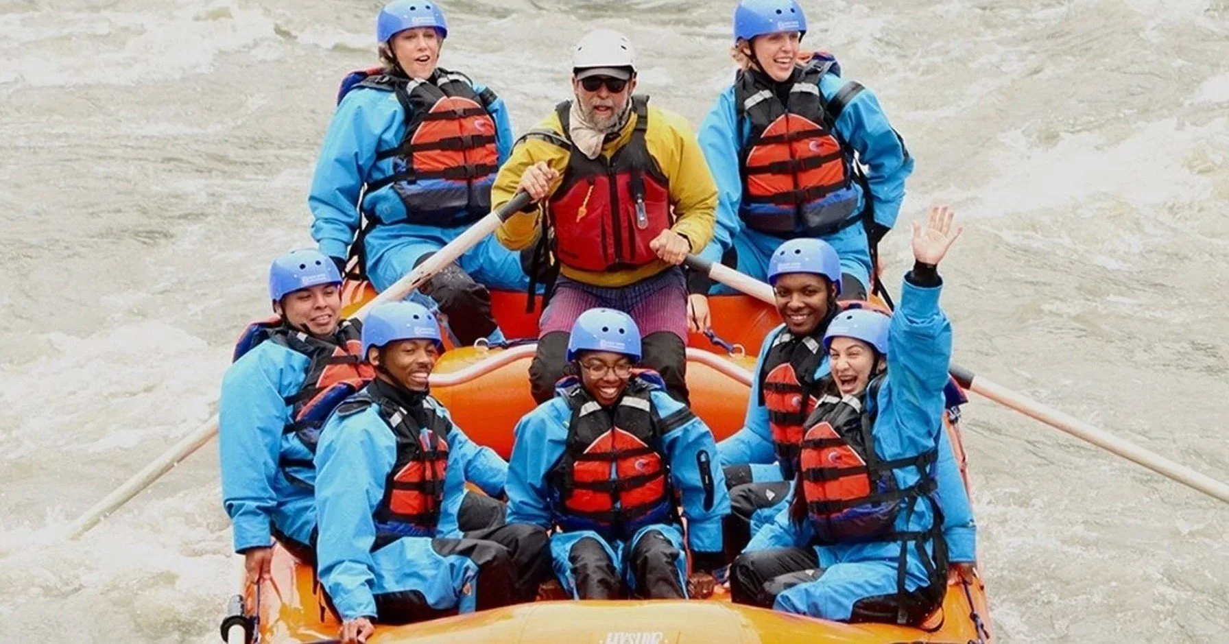 Group of people whitewater rafting in a river, wearing blue helmets and life jackets, with one person in the middle holding a paddle and smiling, surrounded by six others also holding paddles and smiling.