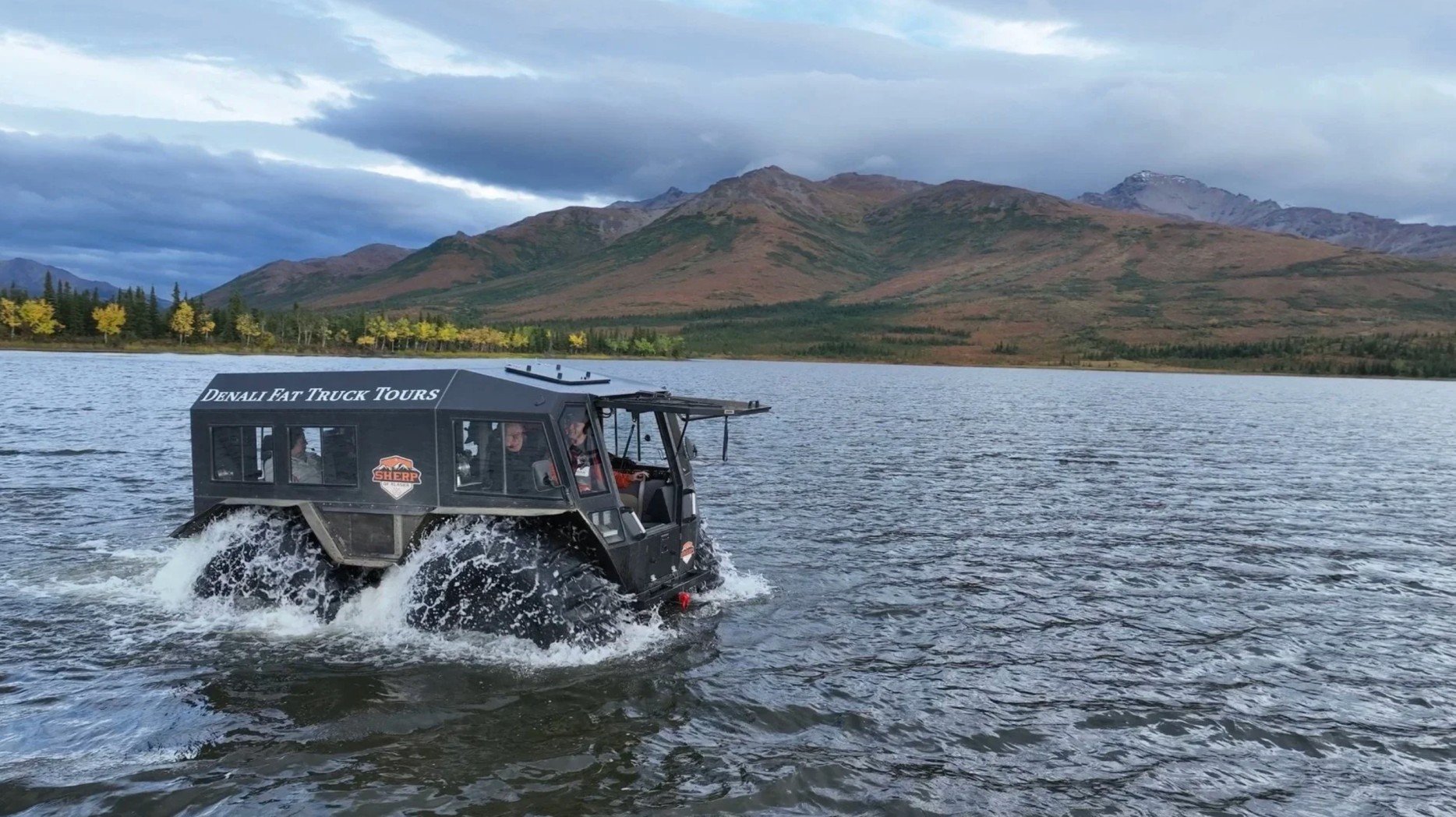 A black Polaris off-road vehicle with a canopy labeled 'Denali Fat Truck Tours' is driving through a body of water, creating splashes, with a mountain range and cloudy sky in the background.