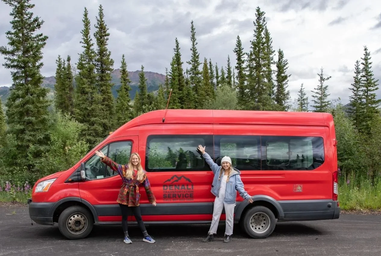 Two women standing in front of a red van with a wooded landscape and cloudy sky in the background. They are smiling and waving at the camera.