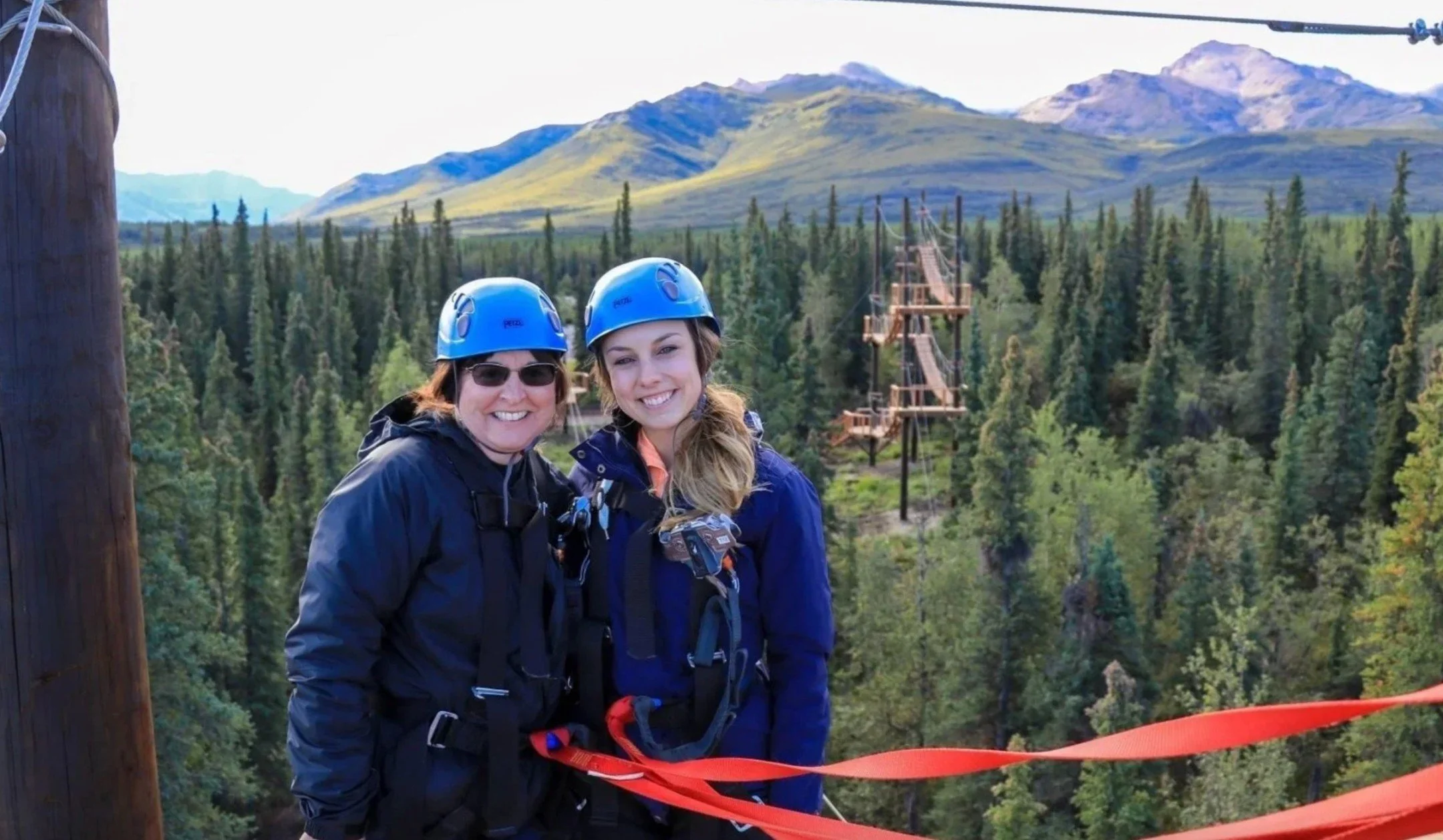 Two women wearing blue helmets and outdoor jackets smiling at the camera, with a mountain landscape and a wooden zip line in the background.