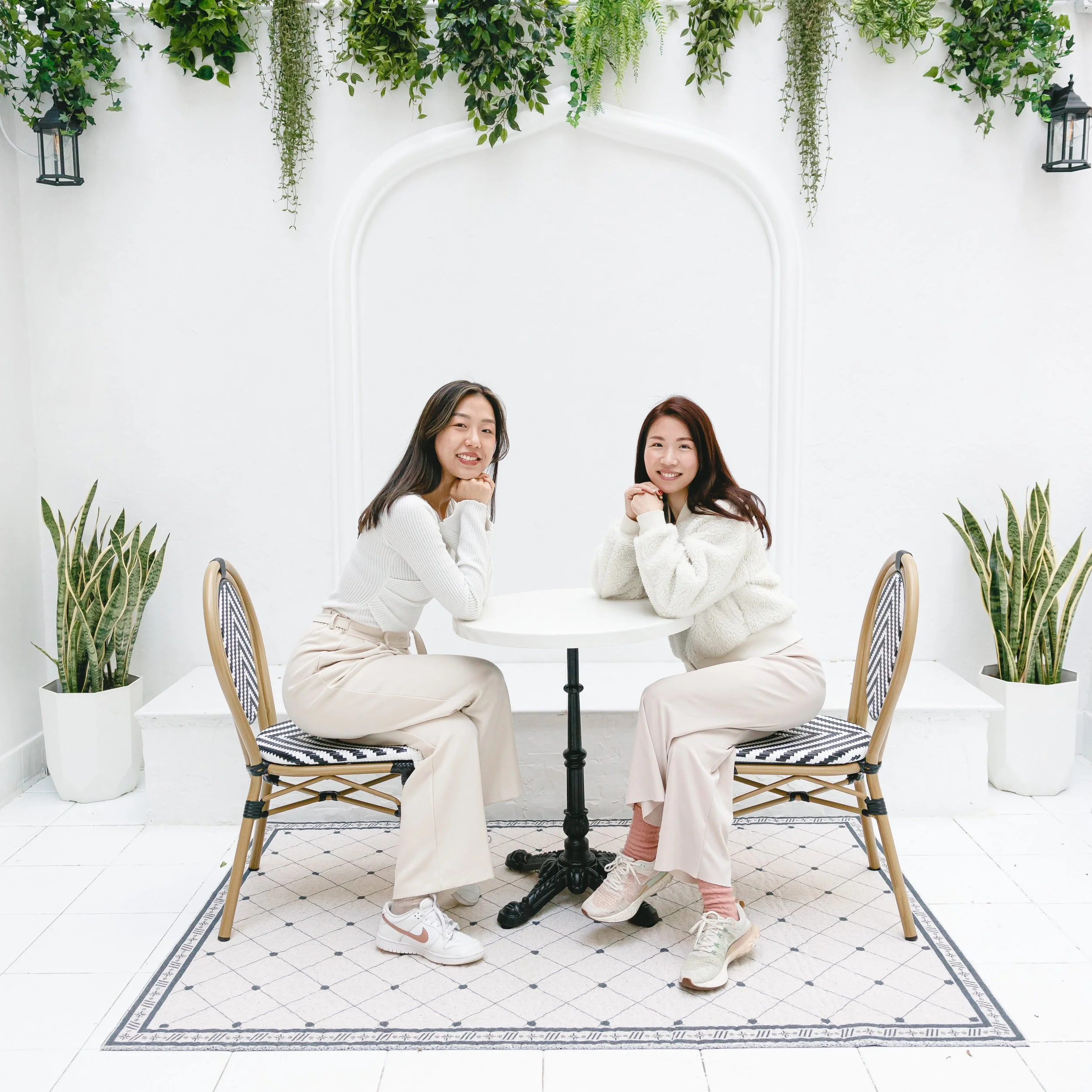Two women sitting at a white round table in a bright, minimalist indoor setting with white walls, green plants, and hanging lights, smiling and resting their heads on their hands.