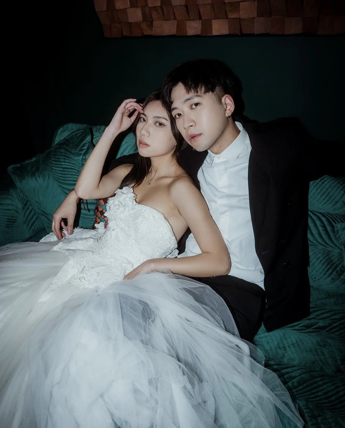 A young woman in a white wedding dress and a young man in a black suit are sitting on a green velvet couch, looking at the camera in a softly lit room.