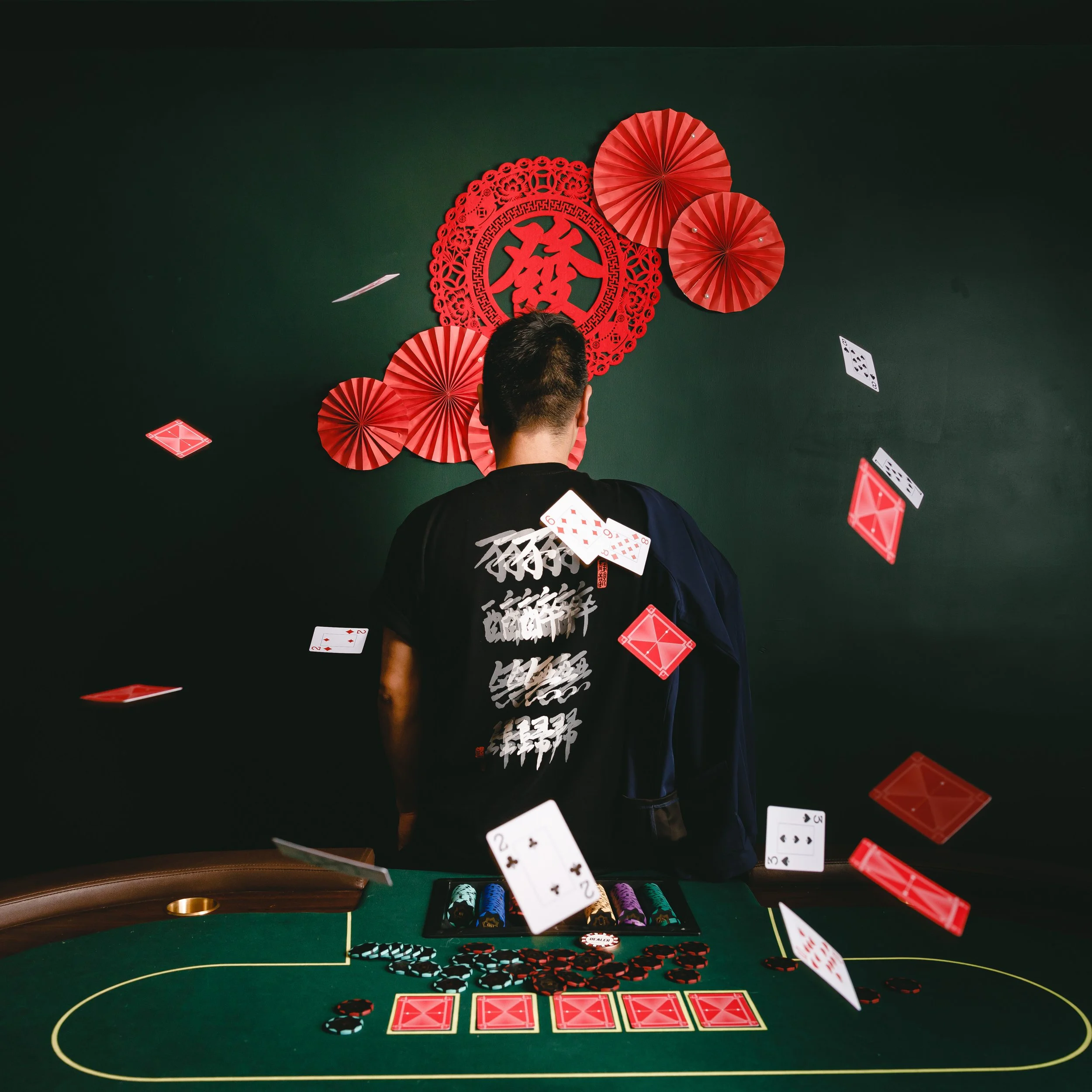 A man facing away standing at a poker table with playing cards and poker chips, with red paper decorations on the dark wall behind him, including a large Chinese character for good fortune.