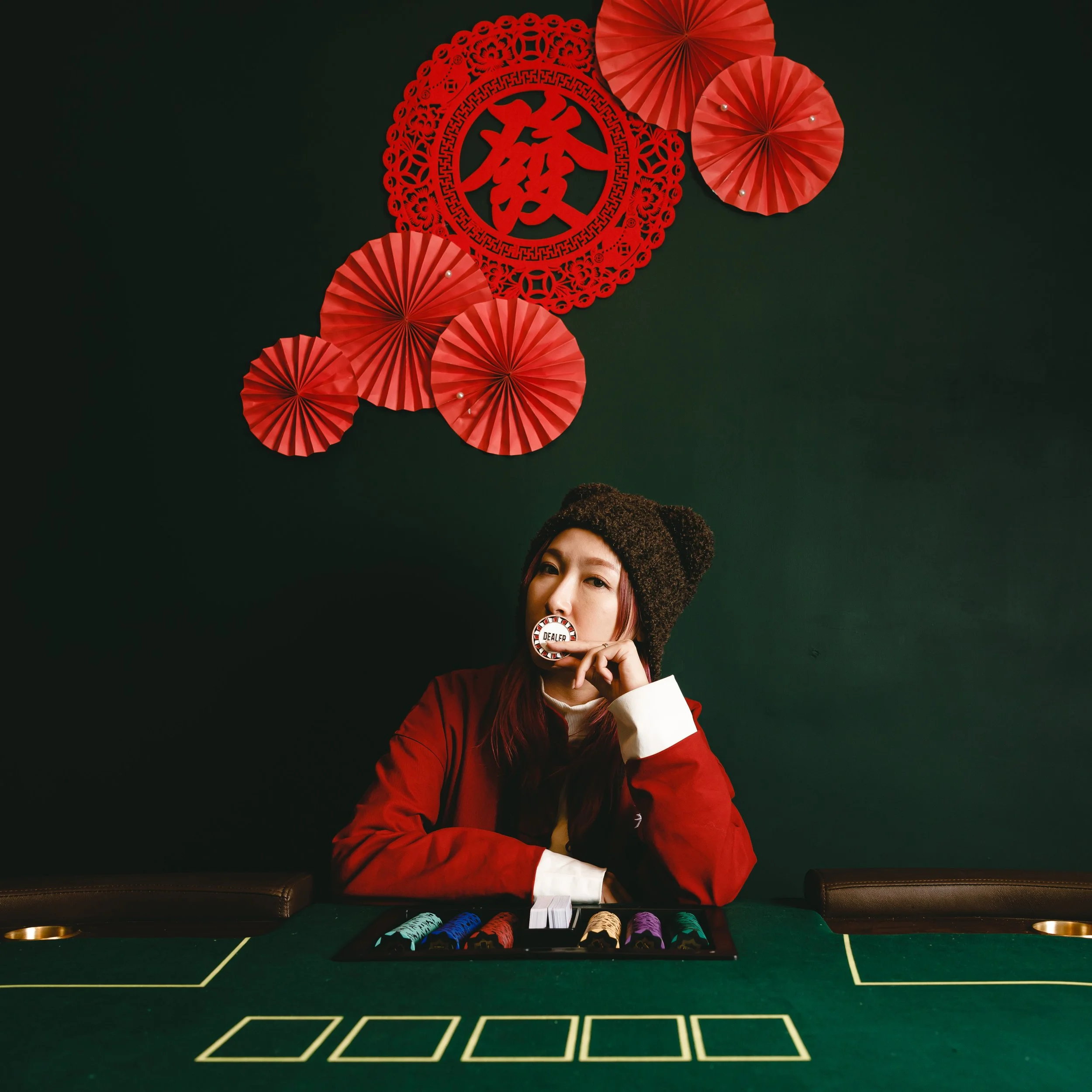 A woman with red hair wearing a red jacket and a brown knit hat sitting at a green poker table with gambling chips. She is holding a poker chip near her mouth and looking at the camera. Behind her, there are red paper fan decorations and a red paper 