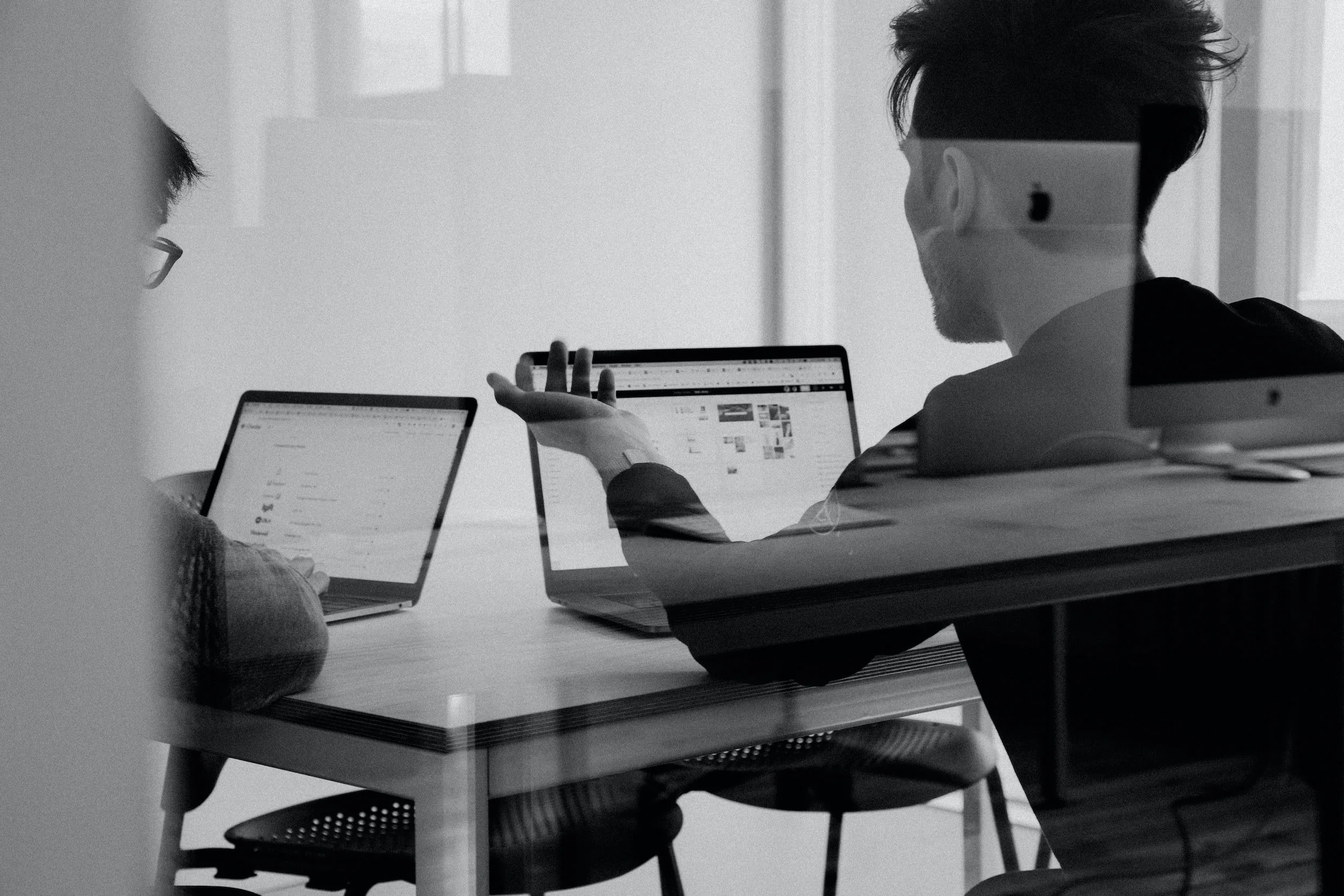 Two people sitting at a desk, using laptops in an office setting.