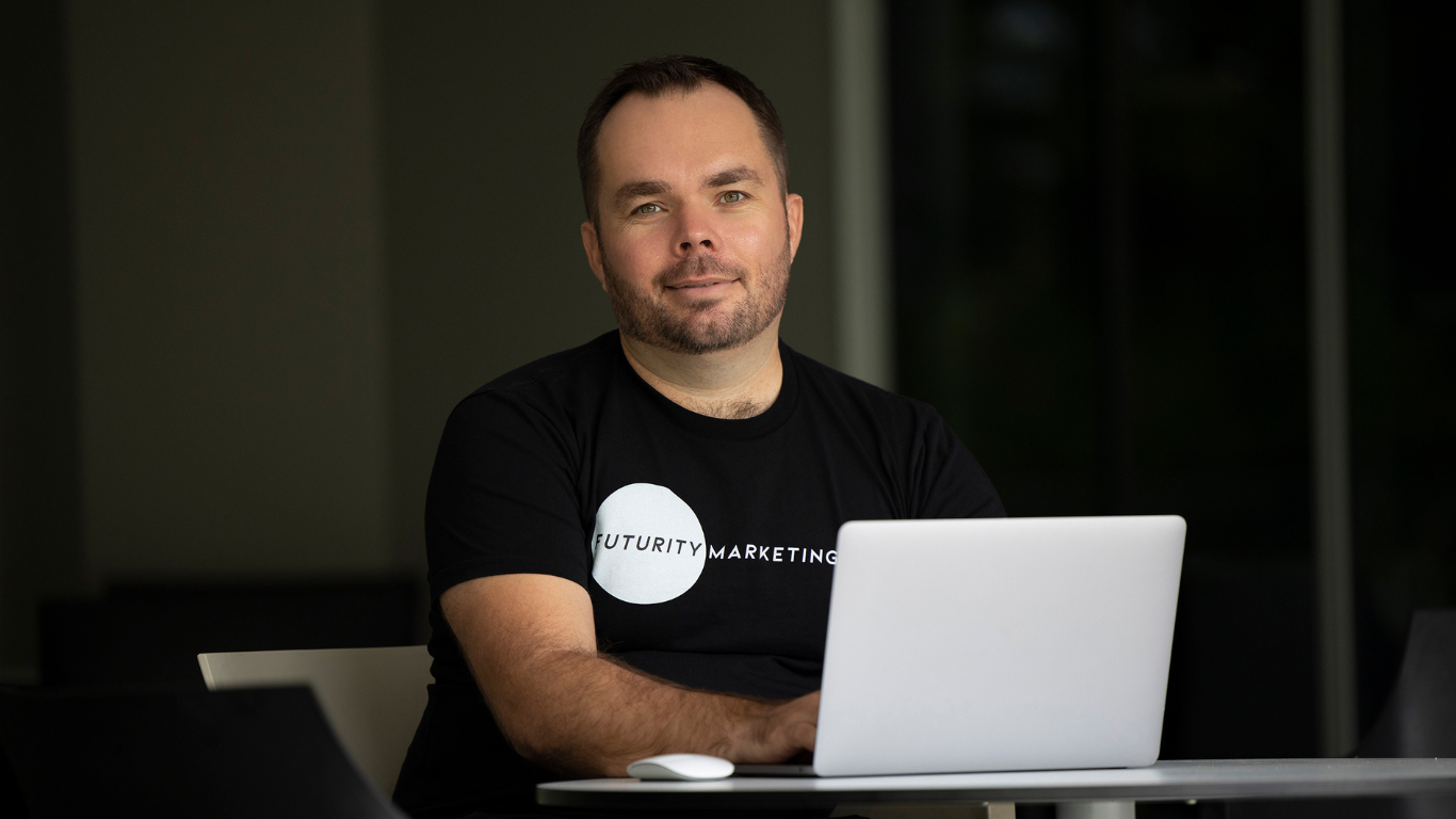 Man wearing a "Futurity Marketing" t-shirt, sitting at a table with a laptop.