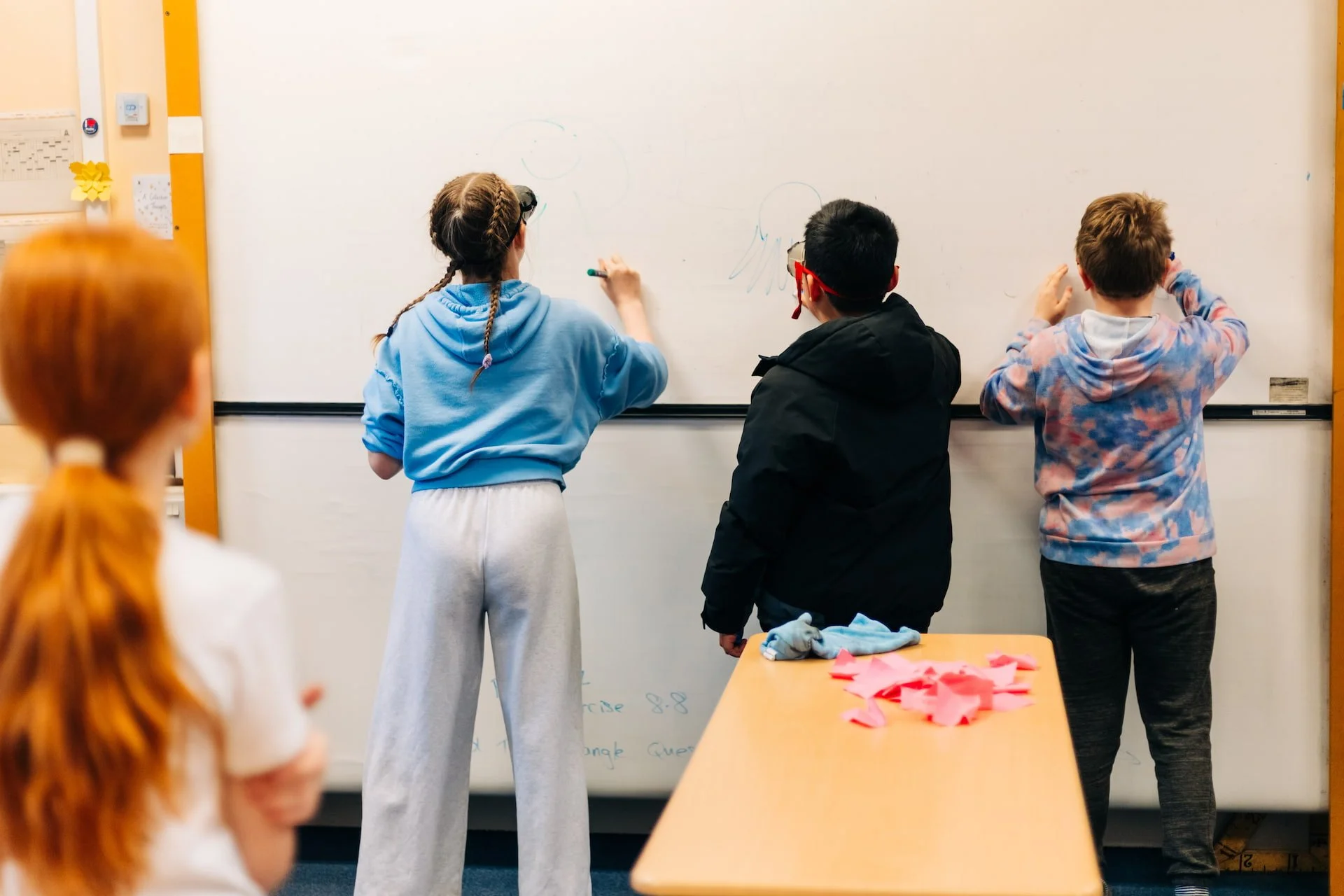 Three children drawing on a whiteboard in a classroom, with a fourth child observing, and pink paper scraps on a table in the foreground.