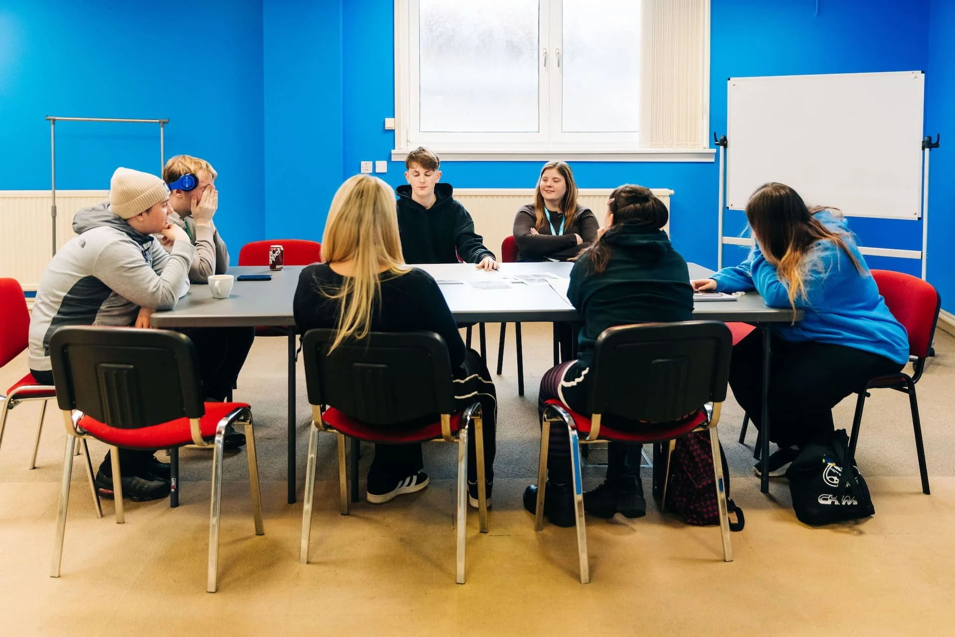 Group of seven young people sitting around a rectangular table in a blue-walled classroom with a whiteboard, engaging in discussion.