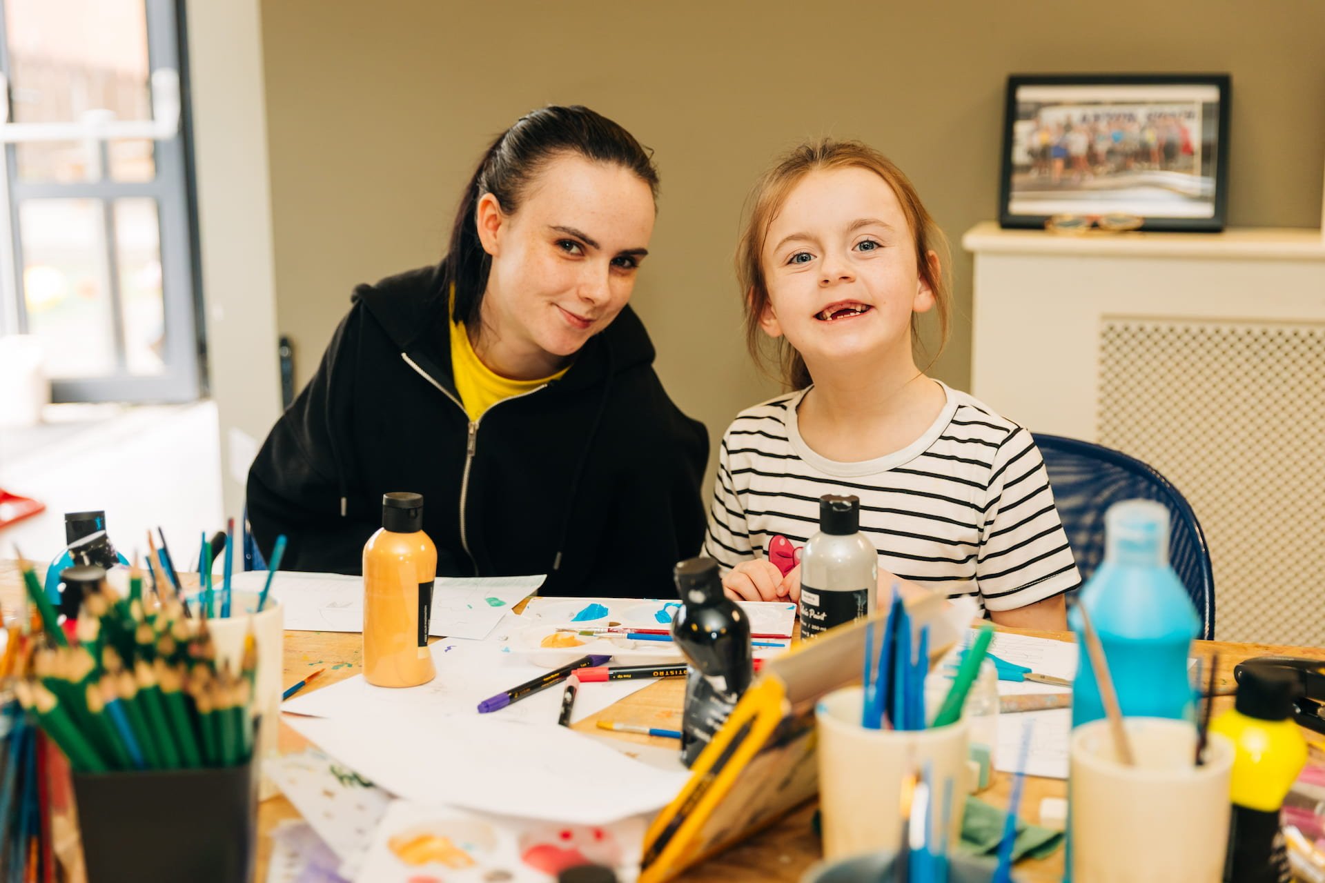 A woman and a young girl sitting at a table filled with art supplies, smiling at the camera.