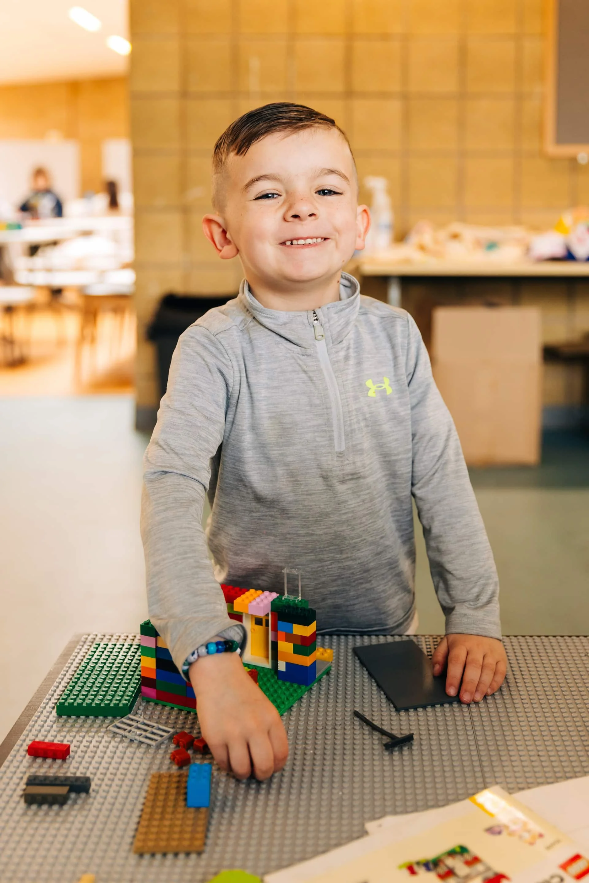 A young boy smiling at a Lego building on a table in a room with a brick wall background.