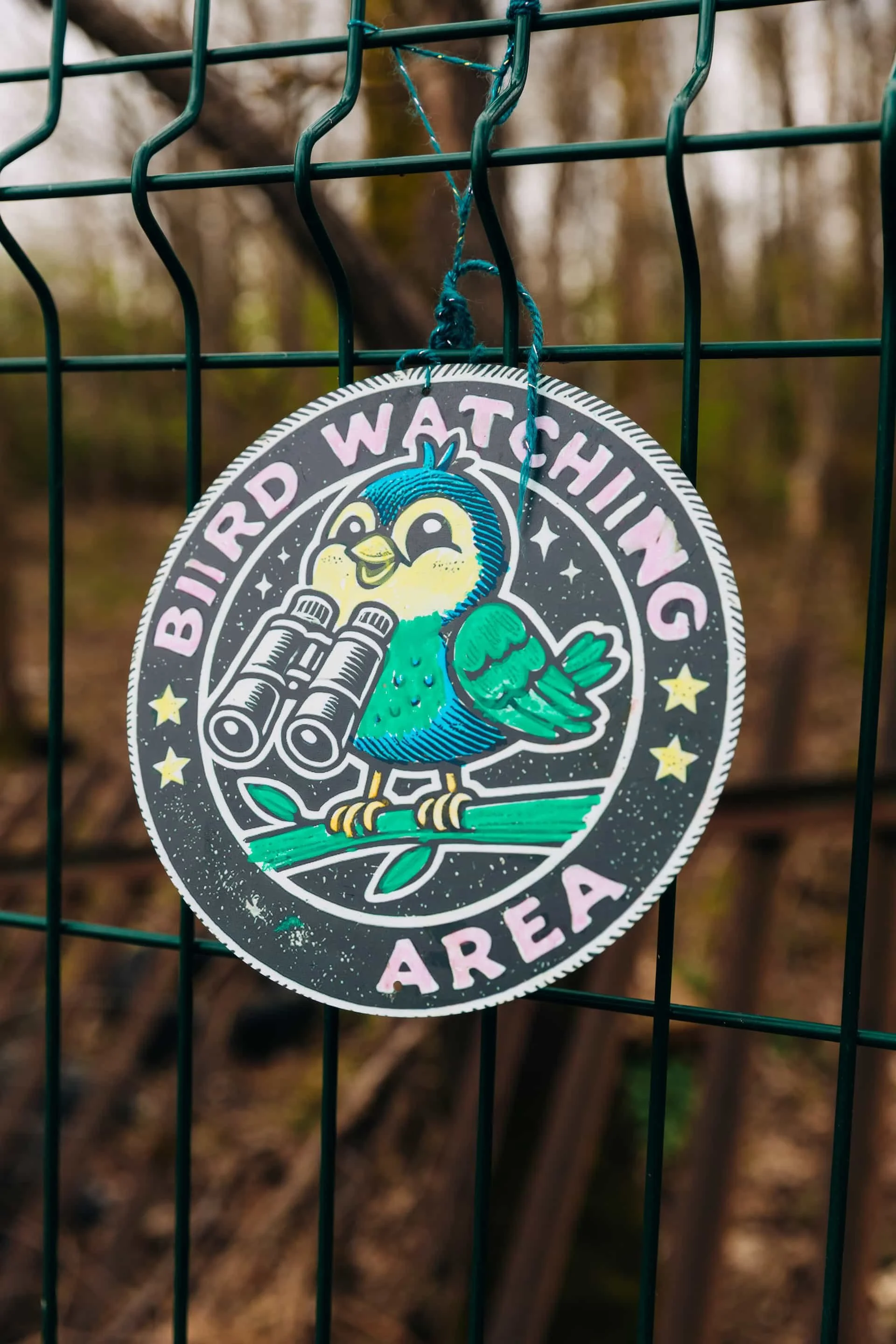 A sign hanging on a green wire fence that says 'Bird Watching Area' with a cartoon bird holding binoculars.
