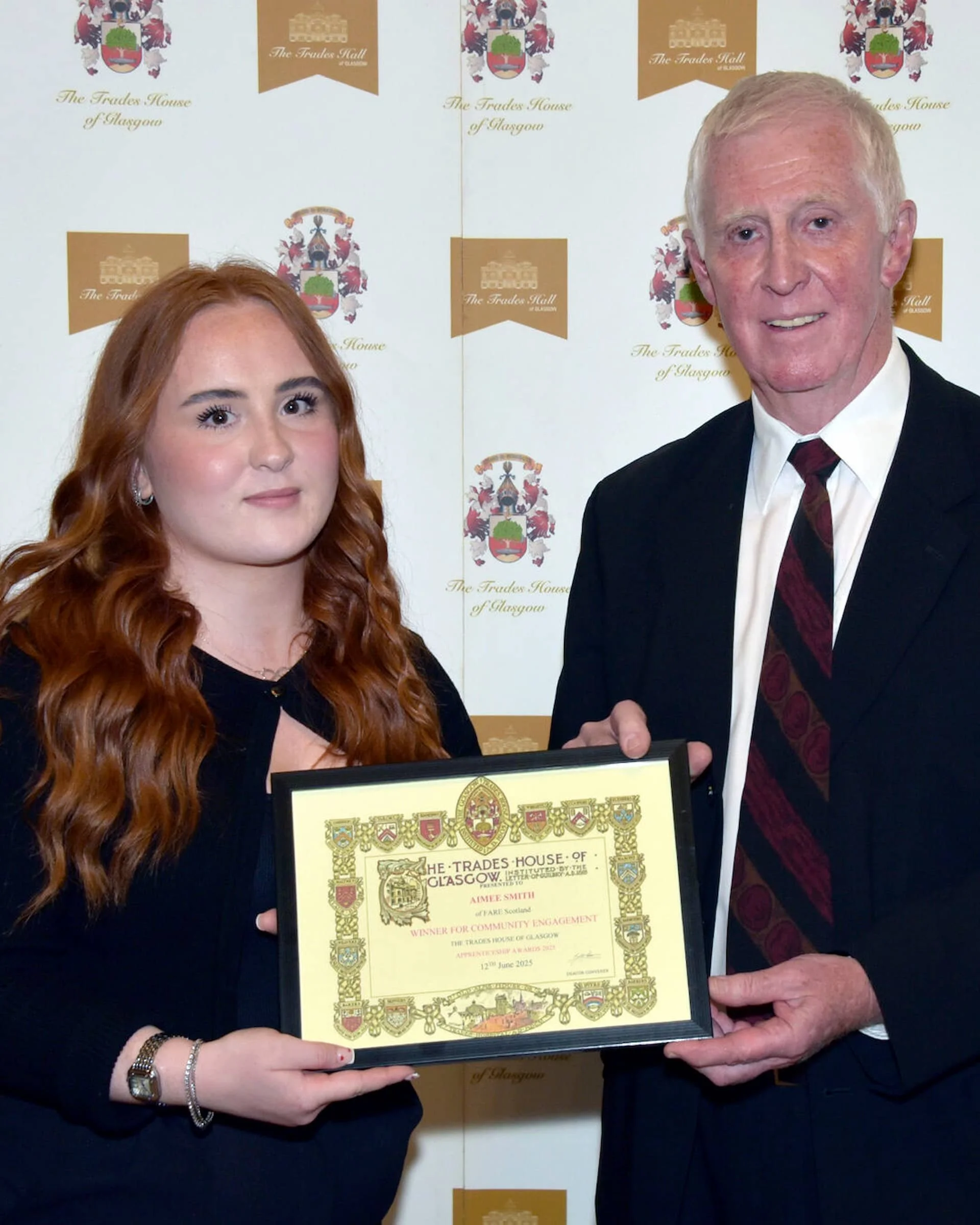 A woman with red hair holding a framed certificate and an older man in a suit shaking hands at an awards ceremony, with a backdrop displaying the logo of The Trades House of Glasgow.