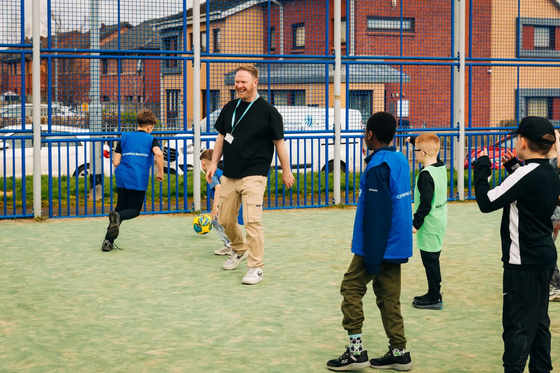 A man with a beard, wearing a black T-shirt and beige pants, is playing soccer with children on a small outdoor field surrounded by a blue fence. Several children are watching or participating, wearing colorful jackets and sportswear. Buildings and cars are visible in the background.