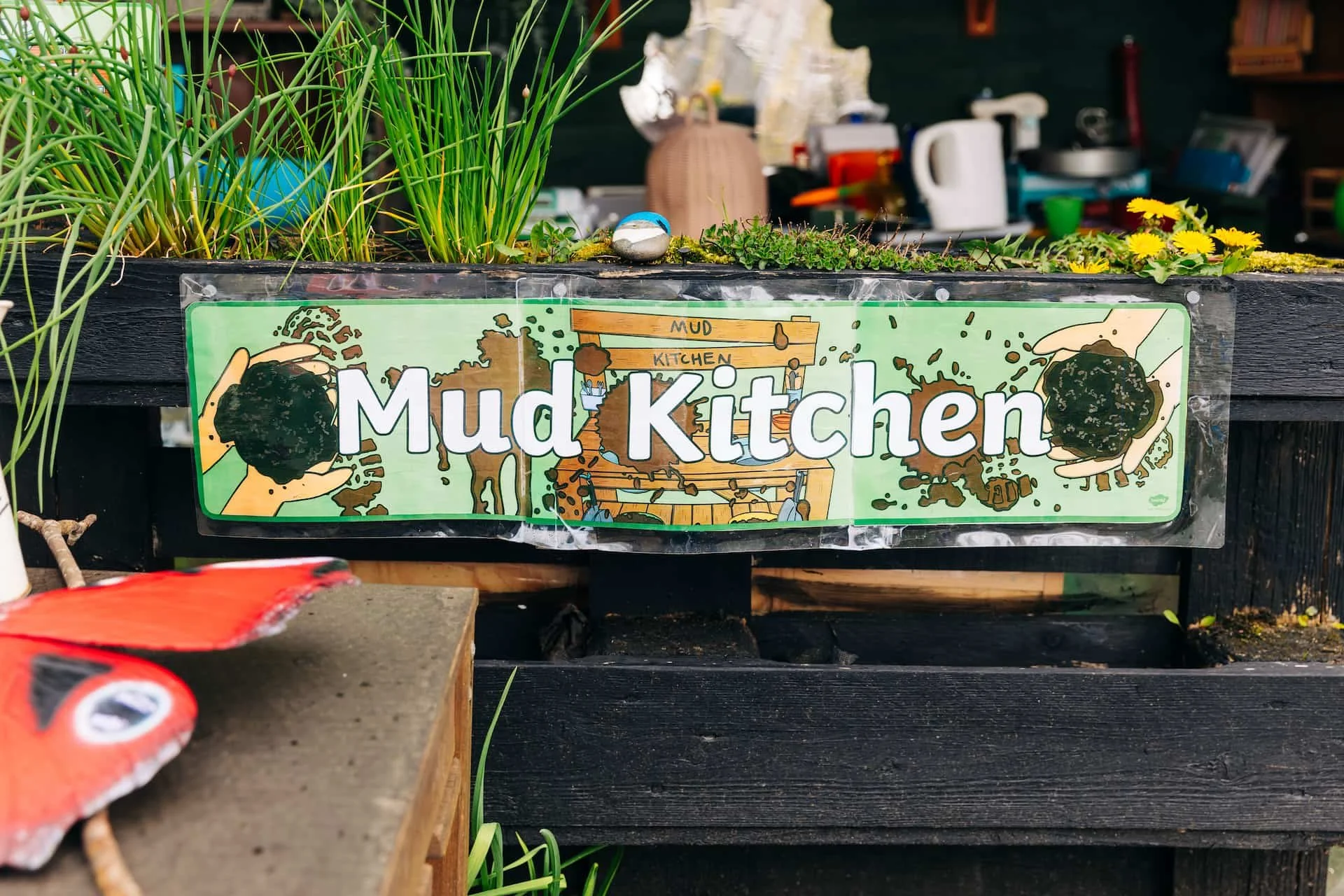 Sign reading 'Mud Kitchen' with illustrations of hands playing with mud and a wooden mud kitchen, surrounded by plants and gardening supplies.