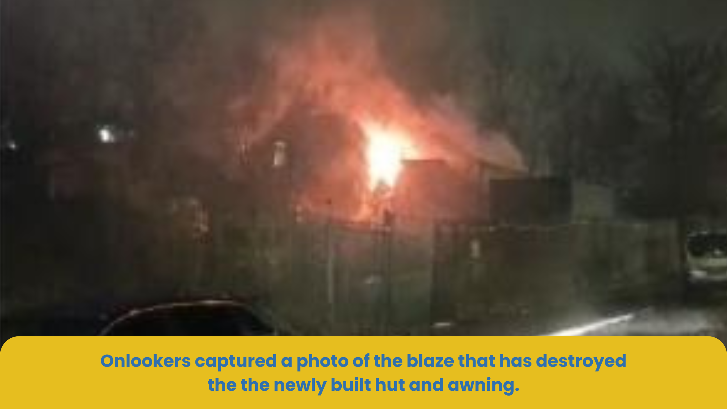 Nighttime photo of a house with flames and smoke coming from the roof, indicating a recent fire. The flames are bright orange and illuminating the dark sky.