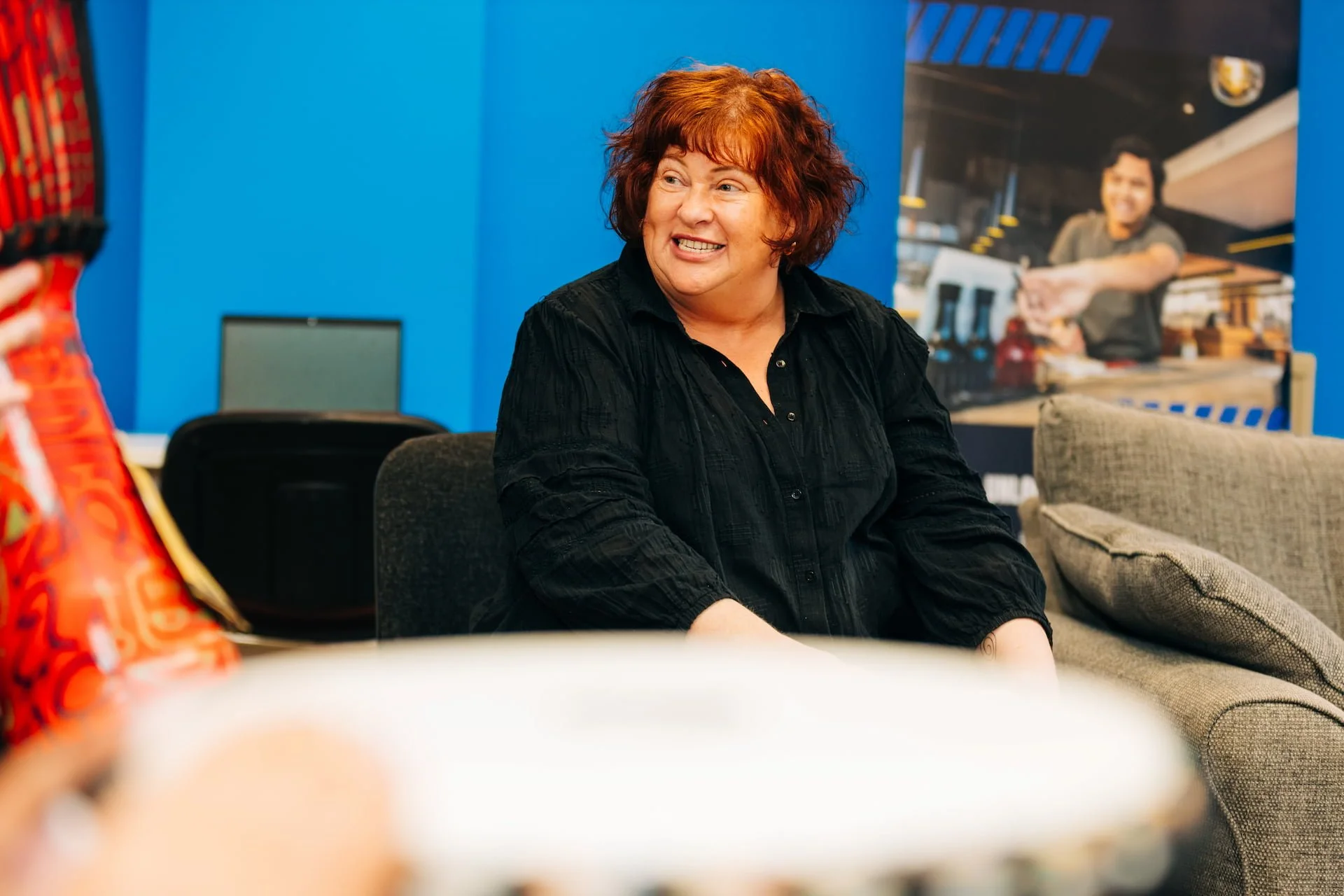 A middle-aged woman with curly red hair, wearing a black shirt, smiling and talking in an office setting with a blue wall and artwork in the background.